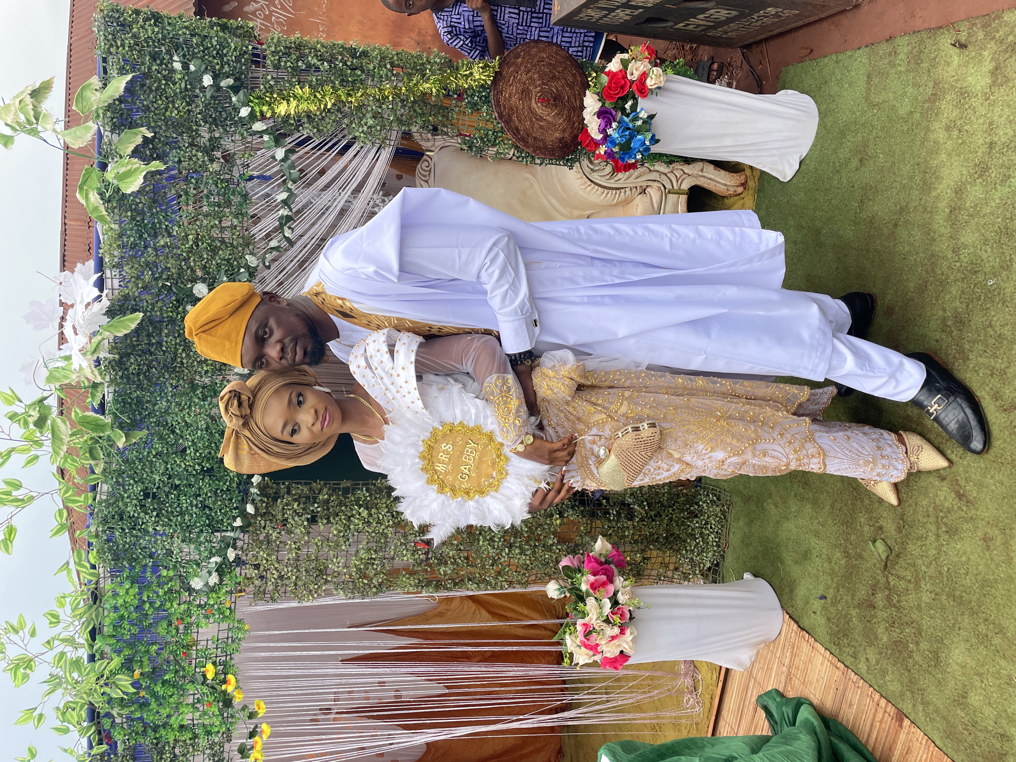 In Ogbe, Nigeria, a couple is photographed in an outdoor setting. The man, wearing a yellow cap and white robe, lies behind the woman. The woman is also in traditional attire and holds a fan that reads "Mrs. Gabby". They are positioned against a backdrop of greenery, white draped fabric, and flower arrangements in white vases. Additional details in the scene include a decorative bench with a woven hat and more flowers. The ground is covered with green turf.