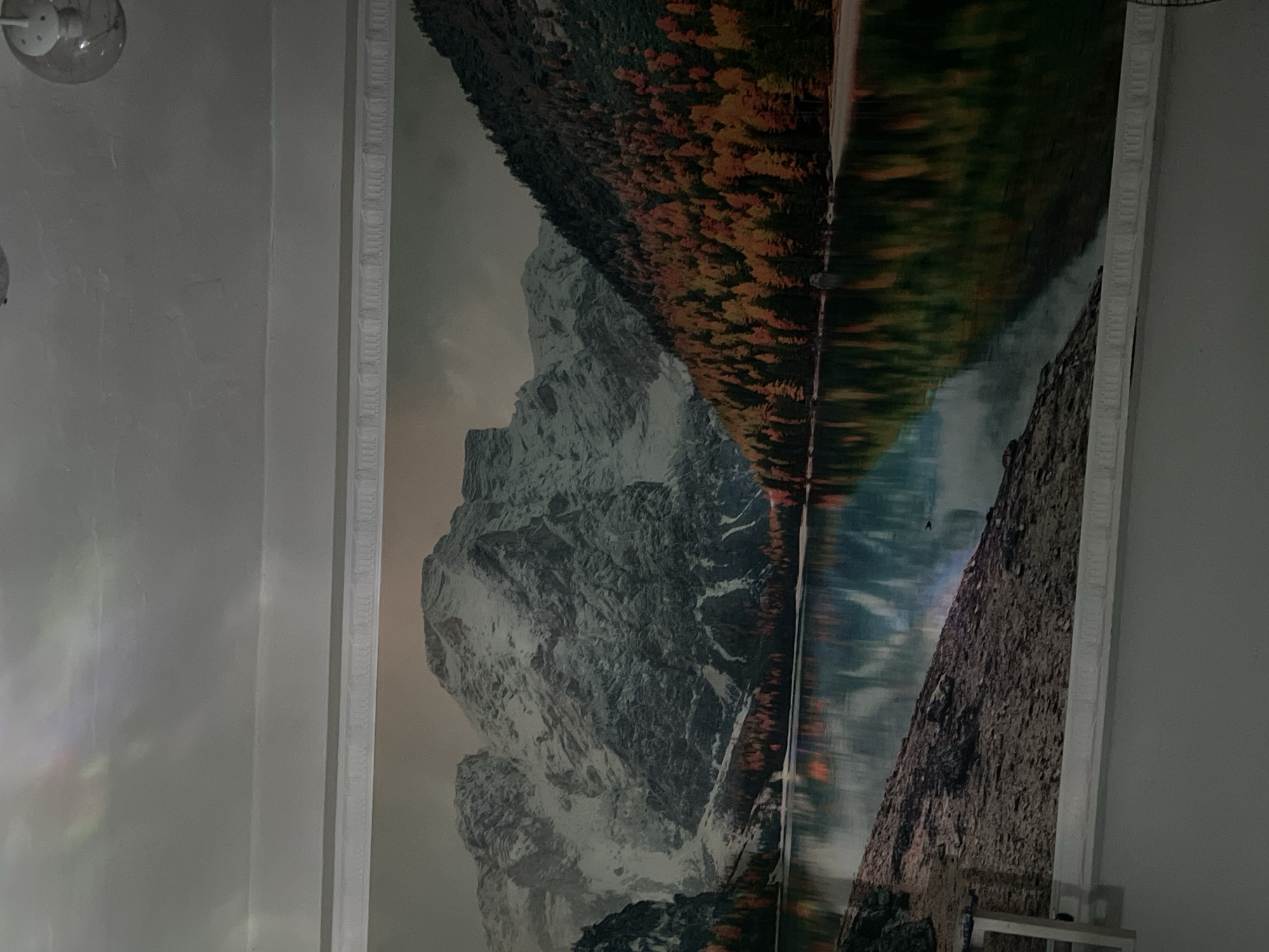 The image shows a large wall mural depicting a scenic mountain landscape with a lake. The mural is framed by decorative molding. In the upper portion of the image, a ceiling light fixture is visible. The mural features snow-capped mountains on the left, a dense forest with autumn foliage on the right, and a calm lake in the foreground reflecting the scenery. The colors in the mural are vibrant, with shades of grey, white, green, orange, and yellow. The overall impression is one of natural beauty and tranquility. There is no indication of the location being in Bishkek City, Kyrgyzstan.