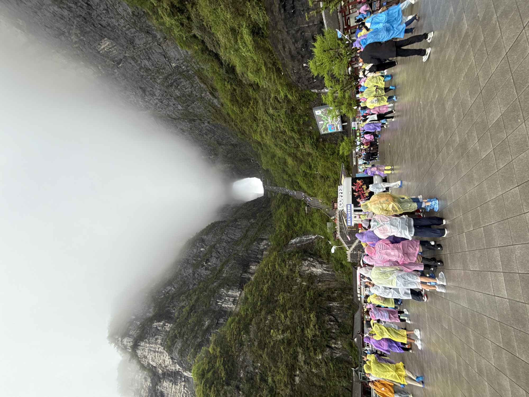 The image captures Tianmen Cave, a natural arch in Tianmen Mountain in the city of Zhangjiajie, Hunan Province, China. The photograph is taken looking towards the cave entrance, which is partially obscured by fog. The mountainside, composed of gray rock, is covered with green vegetation.
In the foreground, people stand on a gray tiled platform. Many are wearing colorful raincoats - shades of yellow, pink, purple, white, and blue. The group seems to be lining up or waiting, possibly for access to a path leading towards the cave.