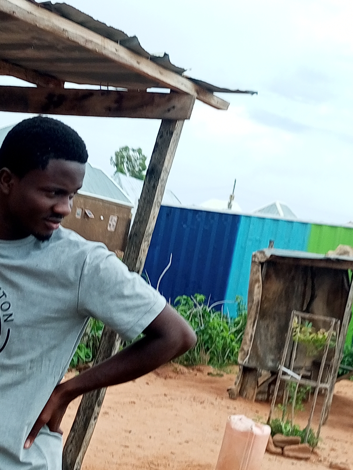 A medium shot captures a young Black man standing outdoors, his body angled slightly to the left, with his left hand on his hip. He is wearing a light gray t-shirt with faint lettering visible on the left chest. He has short dark hair and a short beard. The background reveals a rustic outdoor setting. To the left, a makeshift wooden structure with a corrugated metal roof is visible. Further back, a tall, multi-colored corrugated metal wall, painted in bands of blue, light blue, and green, serves as a backdrop. In the immediate foreground, sandy soil is visible. To the right of the man, a rough wooden structure with an open front and a metal frame containing greenery is present. The sky above is overcast with light clouds. The overall environment suggests a casual, possibly rural or semi-urban setting in Tempure, Nigeria.
