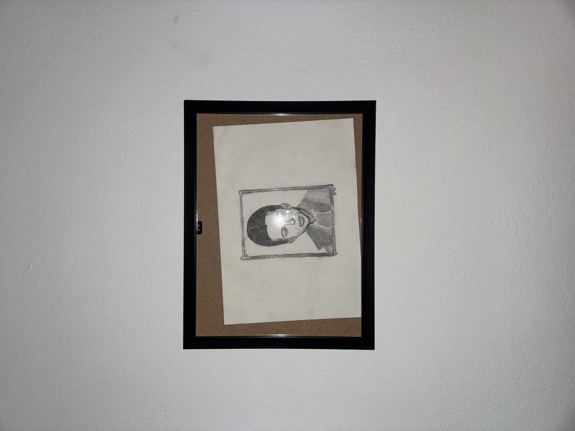 A black-framed picture hangs centered on a white, textured wall, which exhibits minor imperfections and scuffs. Inside the frame, a pencil sketch on light-colored paper is visible, positioned slightly askew, revealing a brown backing board.
The drawing is a stylized portrait, depicting the head and upper torso of a person. The subject's head is tilted, and they appear to be looking slightly upwards and to the viewer's left. The person has dark hair and a neutral to contemplative expression. A distinct, shiny, possibly silver, outline frames the subject within the sketch. Small initials, appearing as "H.H," are visible within this shiny outline on the upper right side of the drawing.
Reflections are apparent on the glass of the picture frame, particularly over the drawing's face, indicating indoor lighting conditions. No specific activity is taking place, and the image does not provide any direct visual cues to a specific geographical location.