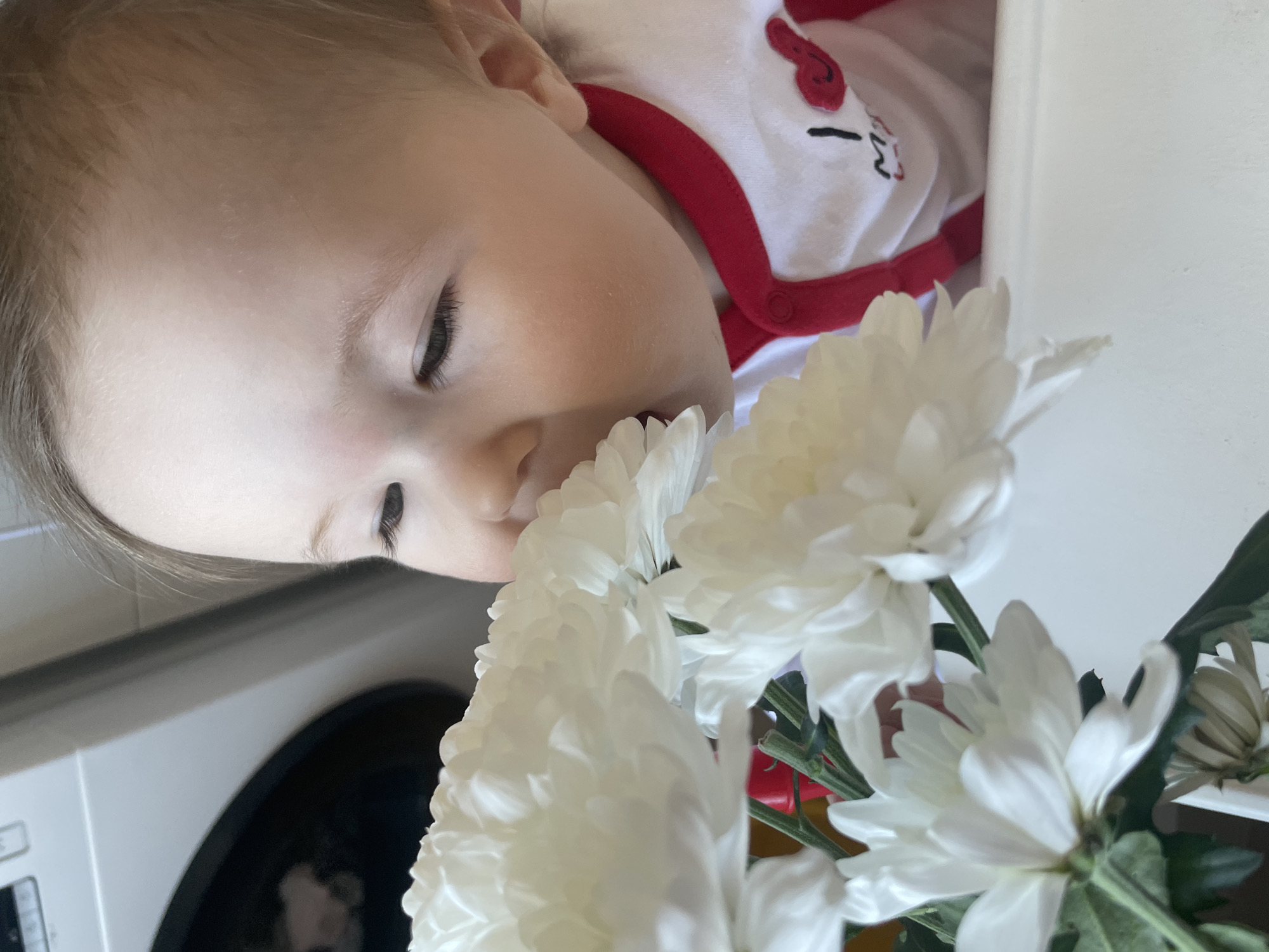 A baby, likely a toddler, is lying on their side, face close to a bouquet of white chrysanthemums. The baby is fair-skinned with closed eyes and dark eyelashes. They are wearing a white outfit with red trim and an illustration of a cartoon cat. The flowers fill the foreground. The background shows part of a white appliance, possibly a washing machine. The location appears to be indoors, likely a private residence, as the prompt notes it is in Chaikovsky, Russia, but there is no verifiable information that confirms that is the case.