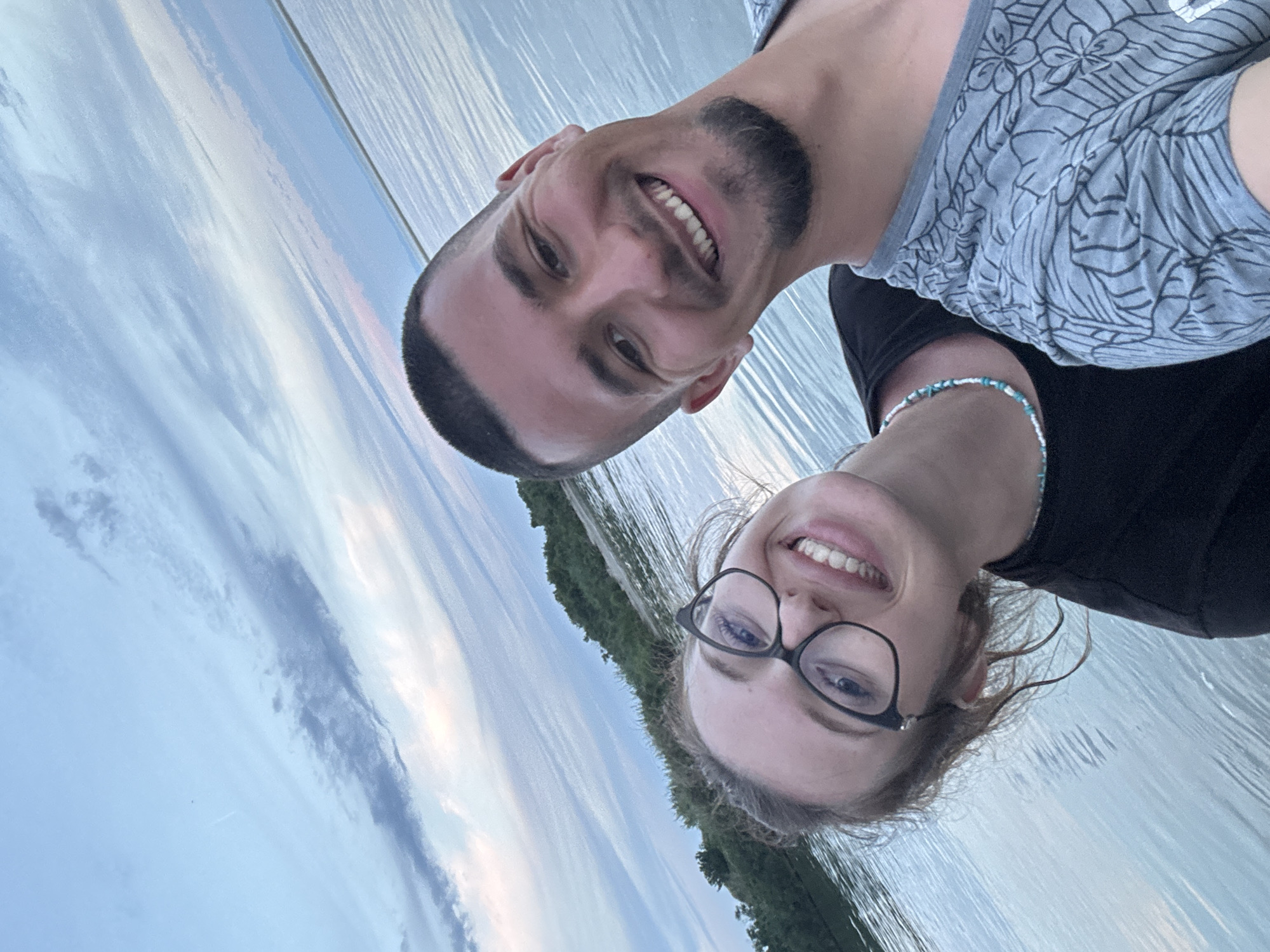 A close-up, low-angle, outdoor, waist-up selfie shows a young adult couple posing by the water at dusk. The man is on the right, with a buzz cut, a short beard, and a grey t-shirt with a leaf pattern. He is smiling widely and looking at the camera. The woman is on the left, wearing glasses and a black t-shirt, and a beaded necklace with blue and white beads. She is also smiling widely and looking at the camera. The water is calm and reflects the sky, which is filled with wispy clouds in shades of blue, grey, and pink. A tree-lined shore is visible in the background. The image is taken in an unknown location in the city of Unknown.