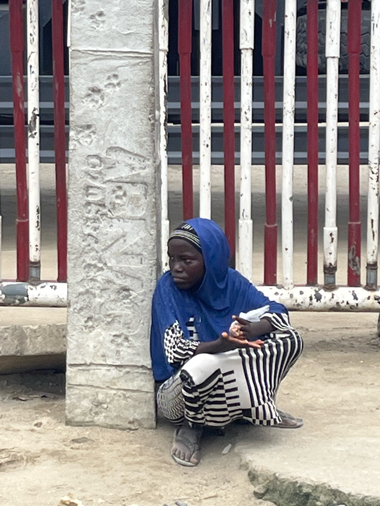A young African person wearing a blue headscarf and a patterned dress is crouched down on the ground next to a concrete pillar. The pillar has textured markings that resemble paw prints and inscribed text. The person is holding a white object in their hands. Behind them, a red and white striped fence stretches across the frame. The ground appears to be dirt or sand. The scene is outdoors, under what seems to be natural light. The location is identified as Ajah, Nigeria.