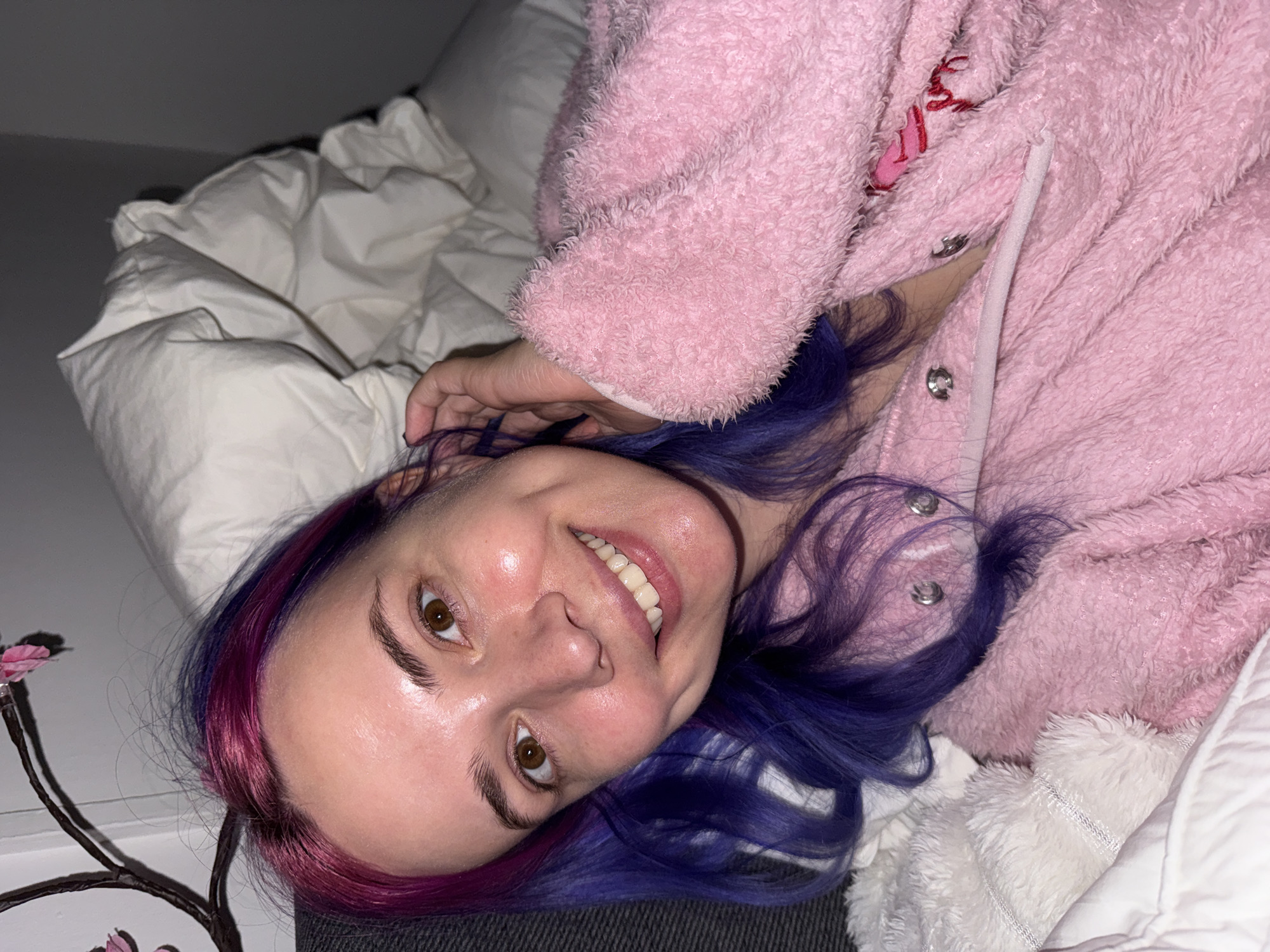 A young woman is prominently featured, lying down and smiling broadly directly at the camera. Her head is tilted, giving the image a dynamic feel. She has light skin, striking brown eyes, and distinctive hair dyed in vibrant shades of purple and magenta, particularly around her face. A small nose piercing is visible.
She is dressed in a cozy, plush, light pink garment, possibly a robe or hooded jacket, which appears to be made of a soft, fluffy material. Silver snap buttons are visible along the front of her attire, and a faint, illegible red embroidery can be seen near the neckline.
The setting is indoors, likely a bedroom, with white bedding, including a duvet and pillow, surrounding her. A dark grey or black surface, possibly a headboard or another blanket, is beneath her head. To her left, a decorative item resembling a branch with dark stems and small pink artificial flowers adds a touch of detail to the scene.
The lighting is soft and warm, suggesting an evening or nighttime atmosphere indoors, contributing to a cozy and intimate mood. The woman's wide smile and relaxed expression convey a sense of happiness and comfort. While not visibly depicted in the image, the provided context indicates this scene takes place in the City of Zagreb, Croatia.