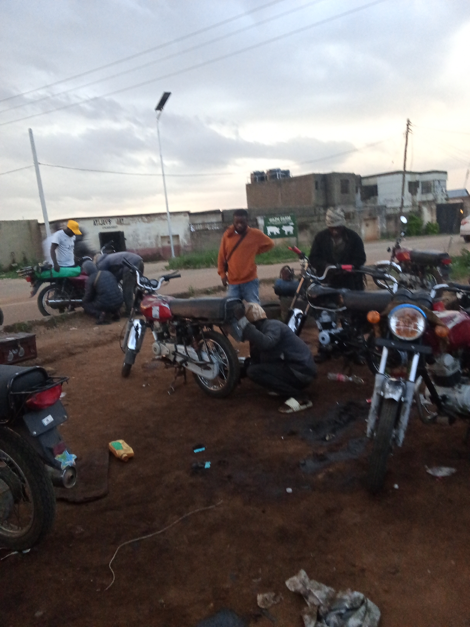 The image depicts an outdoor scene in Barakin Lerawi, Nigeria, likely a roadside workshop or repair area. Several motorcycles are parked, some of which appear to be under repair or maintenance. Three men are visible, actively engaged with the motorcycles. One man, wearing an orange jacket, stands and observes. Another man, in a dark jacket and patterned head covering, is bent over a motorcycle, seemingly working on it. A third individual, wearing a white shirt and a yellow cap, is also near a motorcycle. The ground is dirt, with some scattered debris. In the background, buildings and power lines suggest an urban or semi-urban environment. The sky is overcast.