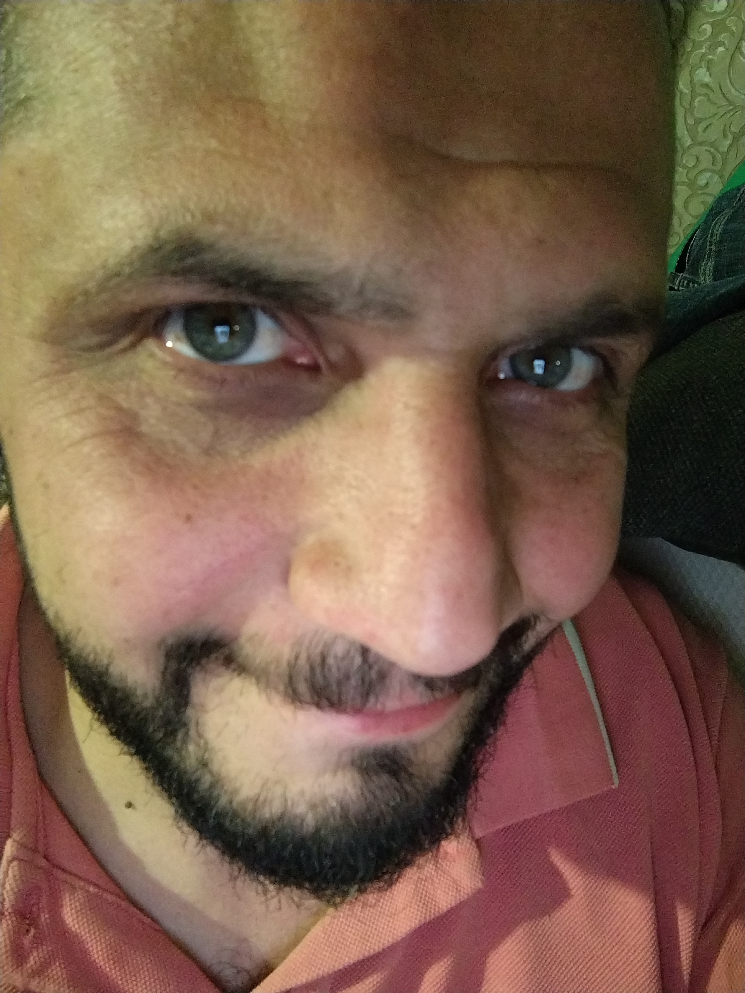 A close-up, eye-level selfie shows a man's face from the nose up, with the camera positioned slightly to his right. The man has medium-toned skin with a light beard and mustache. His eyes are green, and he is wearing a salmon-colored collared shirt. The background is blurred, with a hint of a patterned wallpaper on the left and what appears to be dark fabric on the right. The lighting is warm and appears to come from the front, creating a slight glare in his eyes.