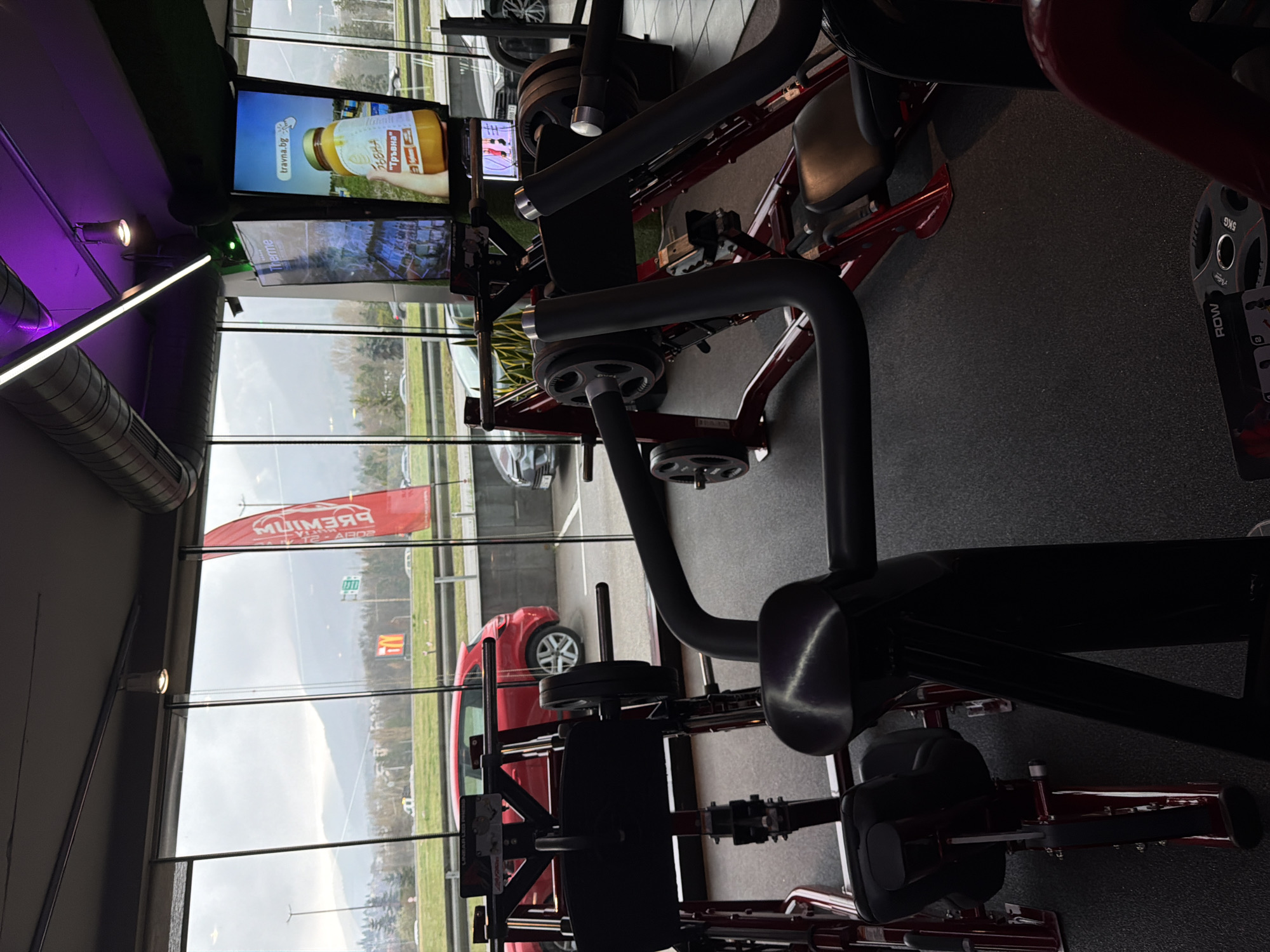 The image presents the interior of a fitness gym in Sofia, Bulgaria, looking outward through large, floor-to-ceiling glass windows. The indoor scene features multiple plate-loaded exercise machines with dark red frames, black upholstery, and dark grey weight plates. A "Linear Leg Press" machine is identifiable in the left foreground. The gym floor is dark, textured rubber. Overhead, the ceiling features exposed silver ductwork, linear white LED lighting, and purple accent lights. Two vertical digital display screens are visible on the right, one showing "Therme" branding and the other displaying an advertisement for "travna.bg" featuring a jar of "Тръвна" honey.
Beyond the windows, an overcast sky hangs over a landscape that identifies the location as Sofia, Bulgaria, due to the visible Vitosha Mountain range in the distance. A multi-lane road or boulevard with vehicular traffic, including a red SUV parked outside, is present in the midground. A yellow McDonald's "M" sign is visible in the distance, alongside a red banner displaying the word "PREMIUM" in reverse. Trees and other distant urban structures complete the exterior view. No individuals are present within the frame.