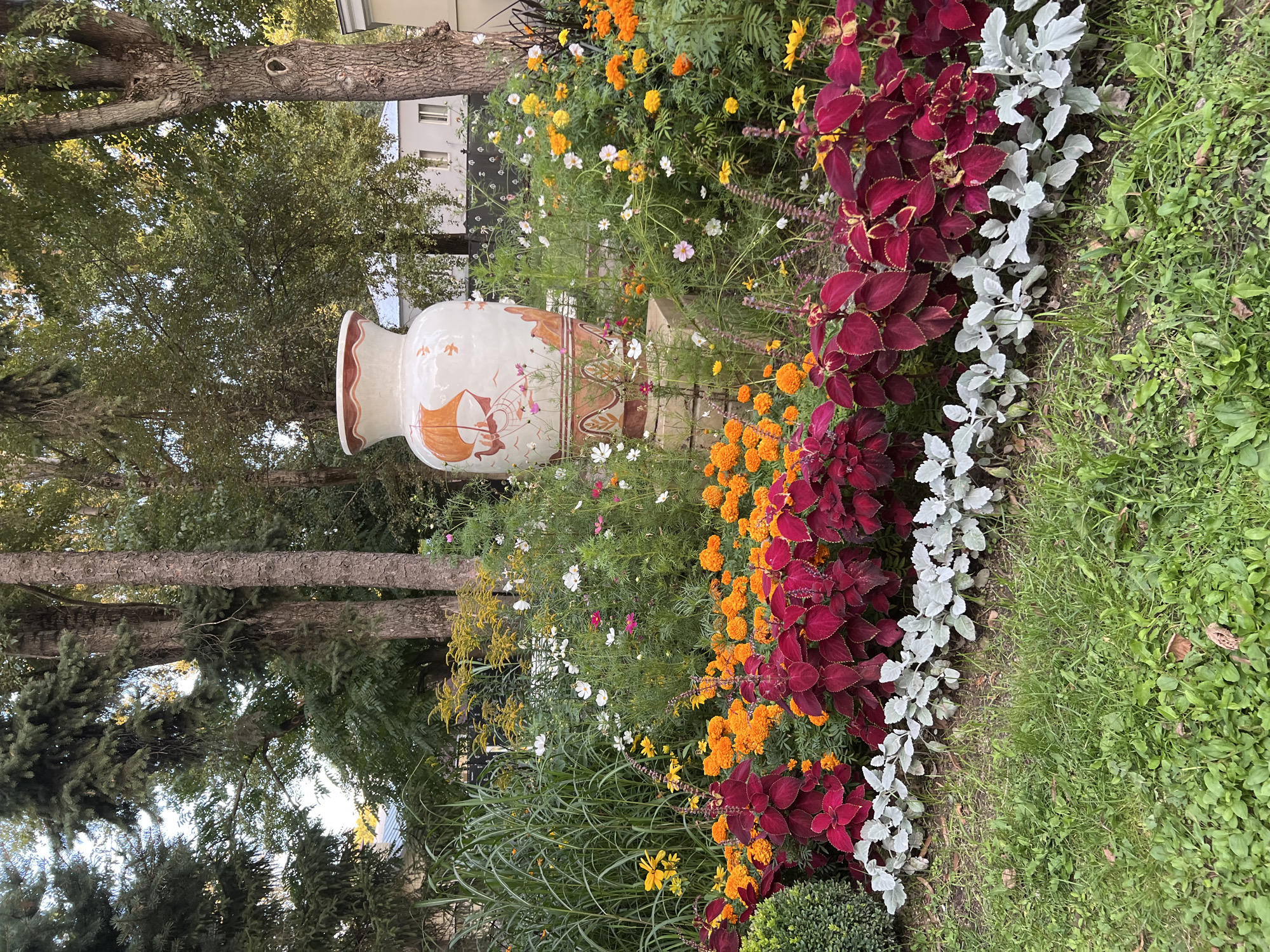 An outdoor, medium-shot photograph displays a flowerbed with a decorative urn as the focal point. The urn, a light-colored ceramic with orange and white painted details depicting a sailing ship and birds, sits on a brick pedestal. The urn is surrounded by a variety of flowers, including vibrant orange marigolds, delicate white and pink cosmos, and yellow flowers. The foreground is filled with lush green grass and bordered by a row of deep red coleus plants and a lower row of silvery-green foliage plants. In the background, tall trees with green foliage provide a natural canopy, and a pale building with windows is visible. The scene appears to be in a park or garden.