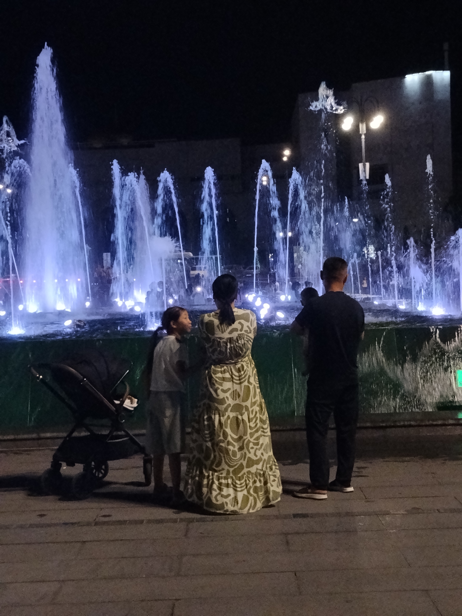 A family of four is standing in front of a brightly lit fountain at night. The father is holding a baby, while the mother and an older child stand beside them. A stroller is parked next to the family. The fountain is illuminated with blue and white lights, creating a dramatic effect against the dark night sky. The scene appears to be in a public park or plaza, possibly in Bishkek City, Kyrgyzstan, given the description provided. The people are dressed casually, and the overall atmosphere seems relaxed and family-oriented.