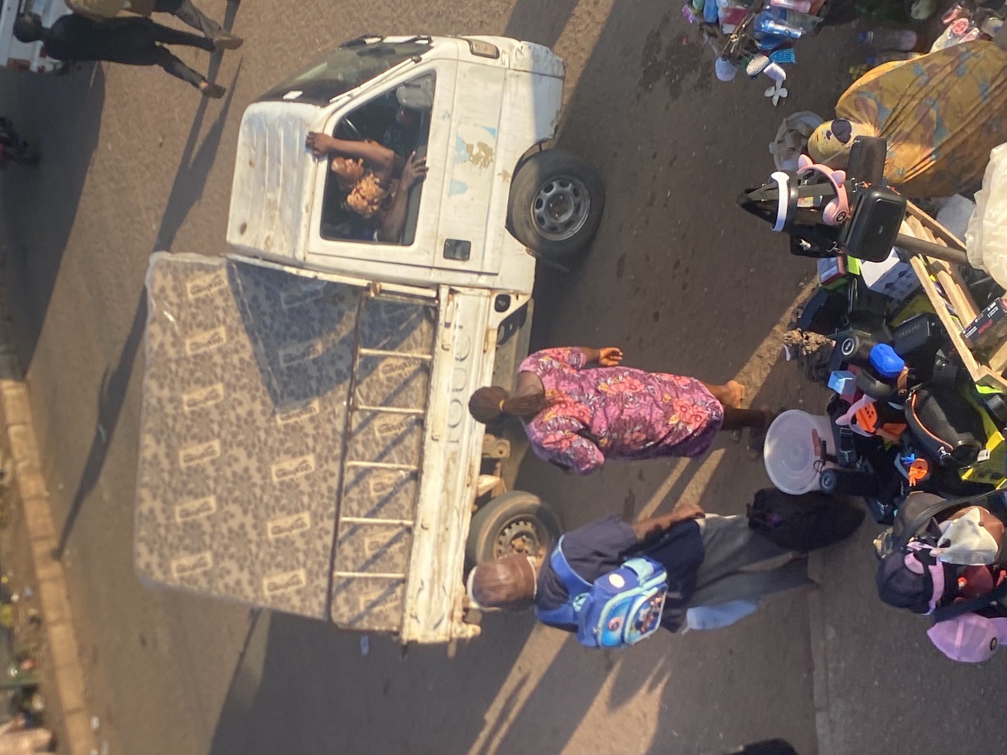 In Bukuru, Nigeria, on a sun-drenched street, a flatbed truck with a white cab is parked. A mattress is loaded onto the truck bed, covered with a patterned tarp. A person with dark skin, wearing a patterned top, is visible through the driver's side window, with their hand resting on the window frame and their head turned to the side. Two individuals stand on the ground near the truck. One, wearing a brown cap and a dark blue shirt with a blue and white backpack, is facing away from the camera. The other, a person with dark skin, wears a vibrant pink and purple floral dress and is facing away from the camera. In the foreground, a collection of items is displayed on a makeshift stand, including backpacks, hats, a white bucket, and various other assorted goods. The background shows a paved road with tire marks and scattered debris.