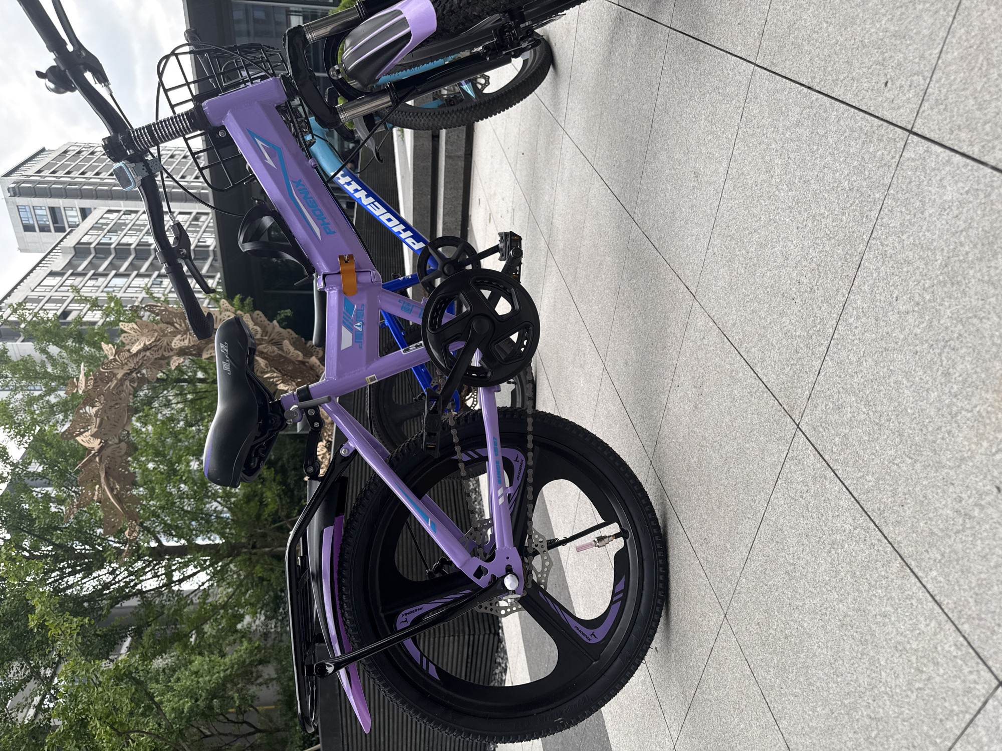 This is a medium shot of a purple and blue folding bicycle parked on a tiled walkway in Yiwu, China. The bicycle is the primary subject, with its frame, wheels, handlebars, and seat clearly visible. The bike has a basket on the front and a rack on the back. The background includes lush green trees and a modern, tall building. The ground is paved with light-colored square tiles, and the lighting suggests it is daytime with diffused sunlight. There are no people or animals in the image.