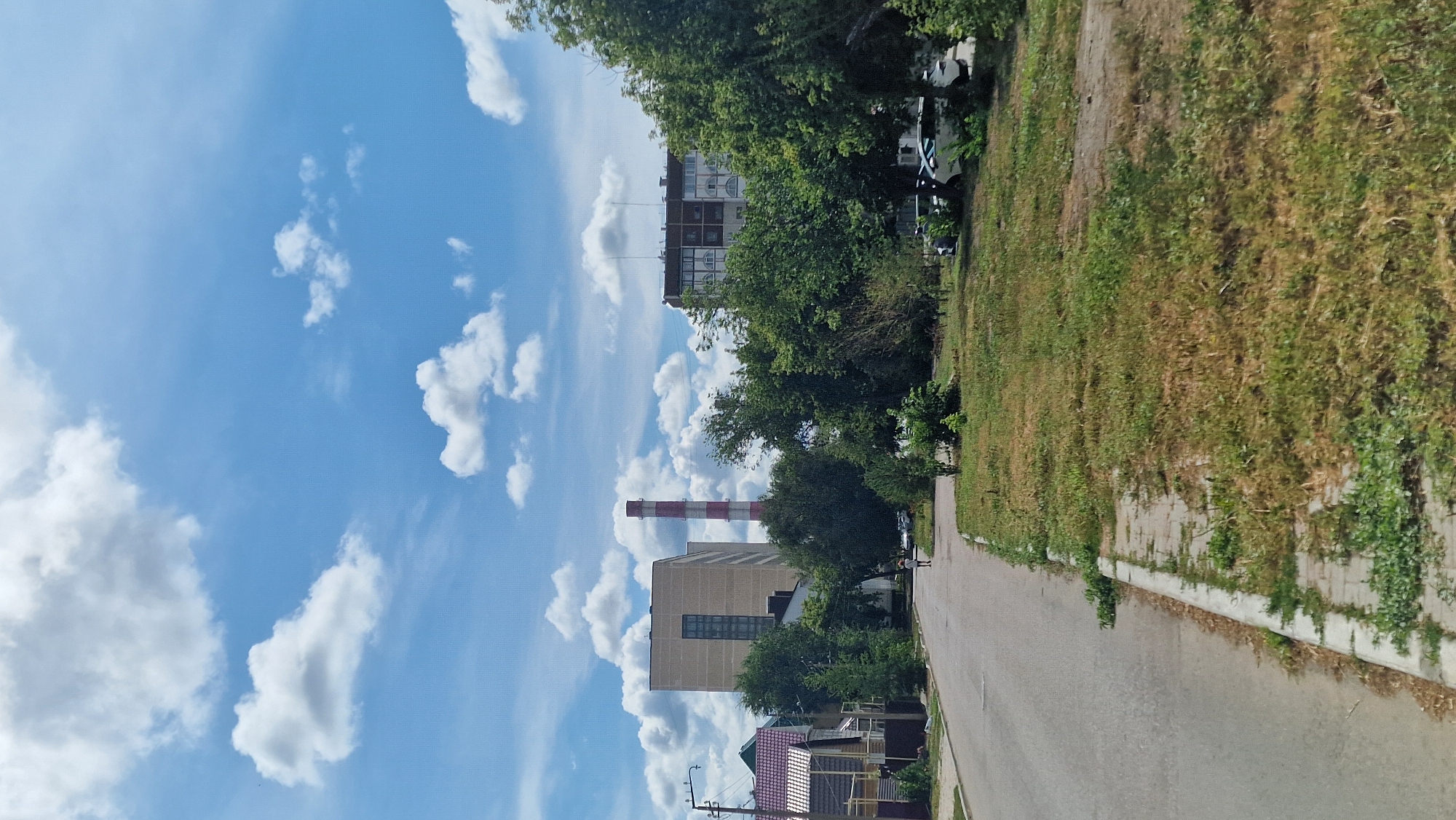 The image depicts a street scene in Kostanay, Kazakhstan. In the foreground, a paved road runs along the left side of the frame, bordered by a grassy area with some sparse vegetation and a concrete curb with paving stones. A lone pedestrian, appearing to be a child, is walking away from the camera down the center of the road.
In the middle ground, several buildings are visible. To the left, a multi-story building with a light-colored facade and a dark vertical window strip is prominent. To its right, a tall, red and white striped industrial chimney stands against the sky. Further to the right, another multi-story apartment building with arched windows is visible. A variety of trees with dense green foliage are interspersed between the buildings and along the edge of the road. Several parked cars are visible in the distance on the right side of the frame.
The background is dominated by a bright blue sky with scattered white, fluffy cumulus clouds. The overall impression is of a typical urban street scene on a clear day.