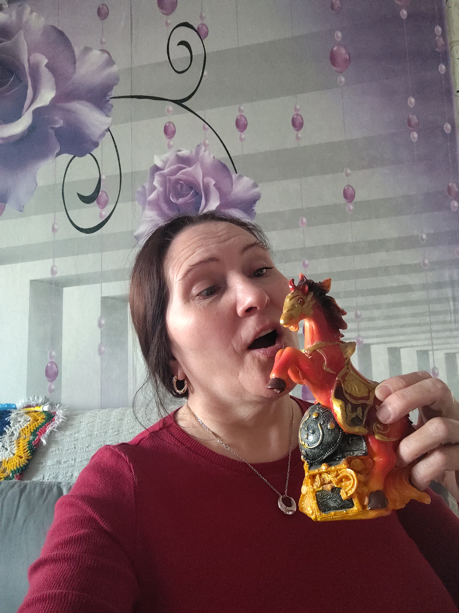 An indoor, eye-level shot captures a middle-aged woman holding a vibrant horse figurine. The woman, with dark brown hair, wears a dark red long-sleeved top and a silver necklace with a circular pendant. A gold hoop earring is visible in her right ear. Her mouth is open, and she looks directly at the horse, suggesting a moment of surprise, playful interaction, or expressive reaction.
She holds a detailed figurine of a rearing horse, predominantly orange-red with elaborate gold accents on its saddle, harness, and base. A black shield-like element is integrated into its pedestal.
The background features a distinctive wallpaper with large, soft purple roses and swirling black vine motifs. Delicate strings of alternating purple and clear beads are also depicted on the wallpaper, set against a subtle horizontally striped grey background. To the left, a light grey couch is visible, topped with a colorful, possibly crocheted or knitted item. The scene suggests a cozy domestic setting, likely within a home in Сусады-Эбалак, Russia. There is no visible text in the image.