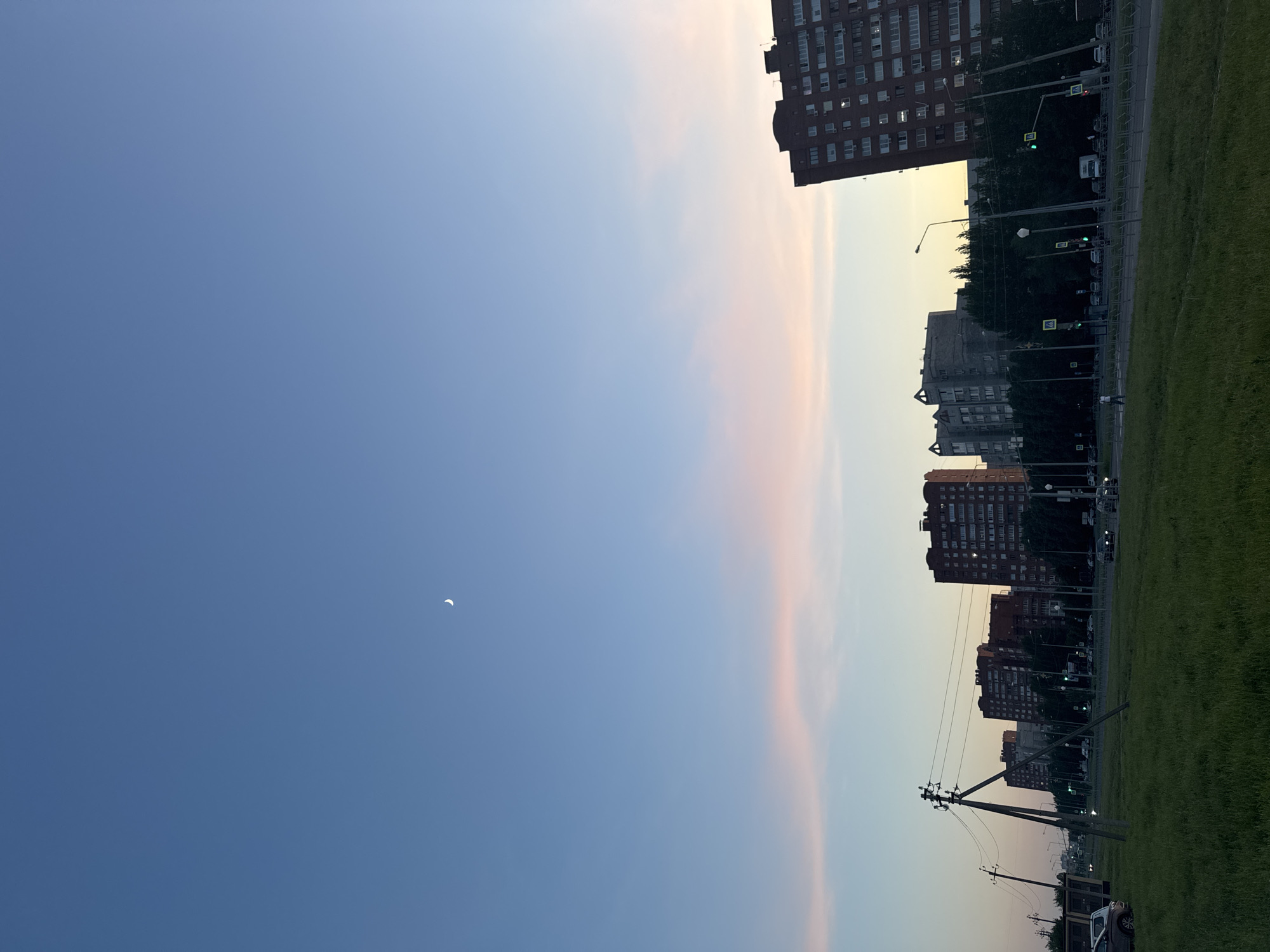 The image depicts a cityscape at dusk in Unknown, Unknown. High-rise brick apartment buildings line the right side of the frame, contrasted against a clear blue sky on the left. A crescent moon is visible in the upper left quadrant. A strip of green lawn and road with traffic lights and pedestrian crossings can be seen in front of the buildings. Utility poles with wires are positioned in the lower right corner, near a parked car. The sky transitions from blue to a pale orange-yellow near the horizon.