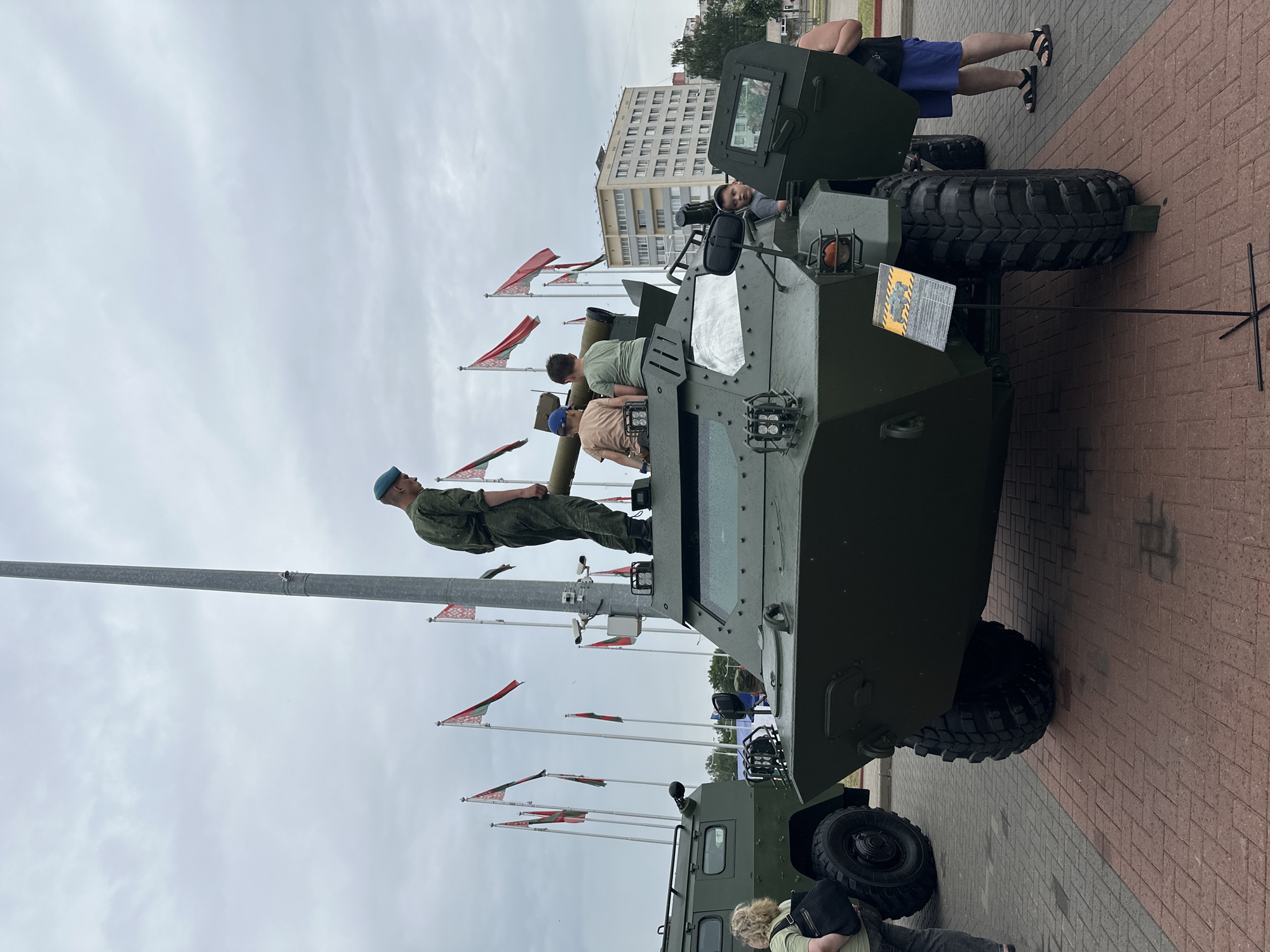 A group of people are gathered around a military vehicle at an outdoor event. In the foreground, a green armored vehicle is on display. A soldier in a green uniform and blue beret stands on top of the vehicle, holding a long tube. Two young boys are also on top of the vehicle, looking forward. Another young boy is inside the vehicle, looking at the camera. To the left, a woman in a light-colored shirt and dark pants stands with a black bag. Behind the armored vehicle, another green military vehicle is visible. Numerous flags are flying in the background. In the far background, a tall building with many windows can be seen. The event appears to be taking place on a paved surface, possibly a city square, with a brick pattern. The sky is overcast. The event is identified as taking place in Minsk, Belarus, through the presence of the national flag.