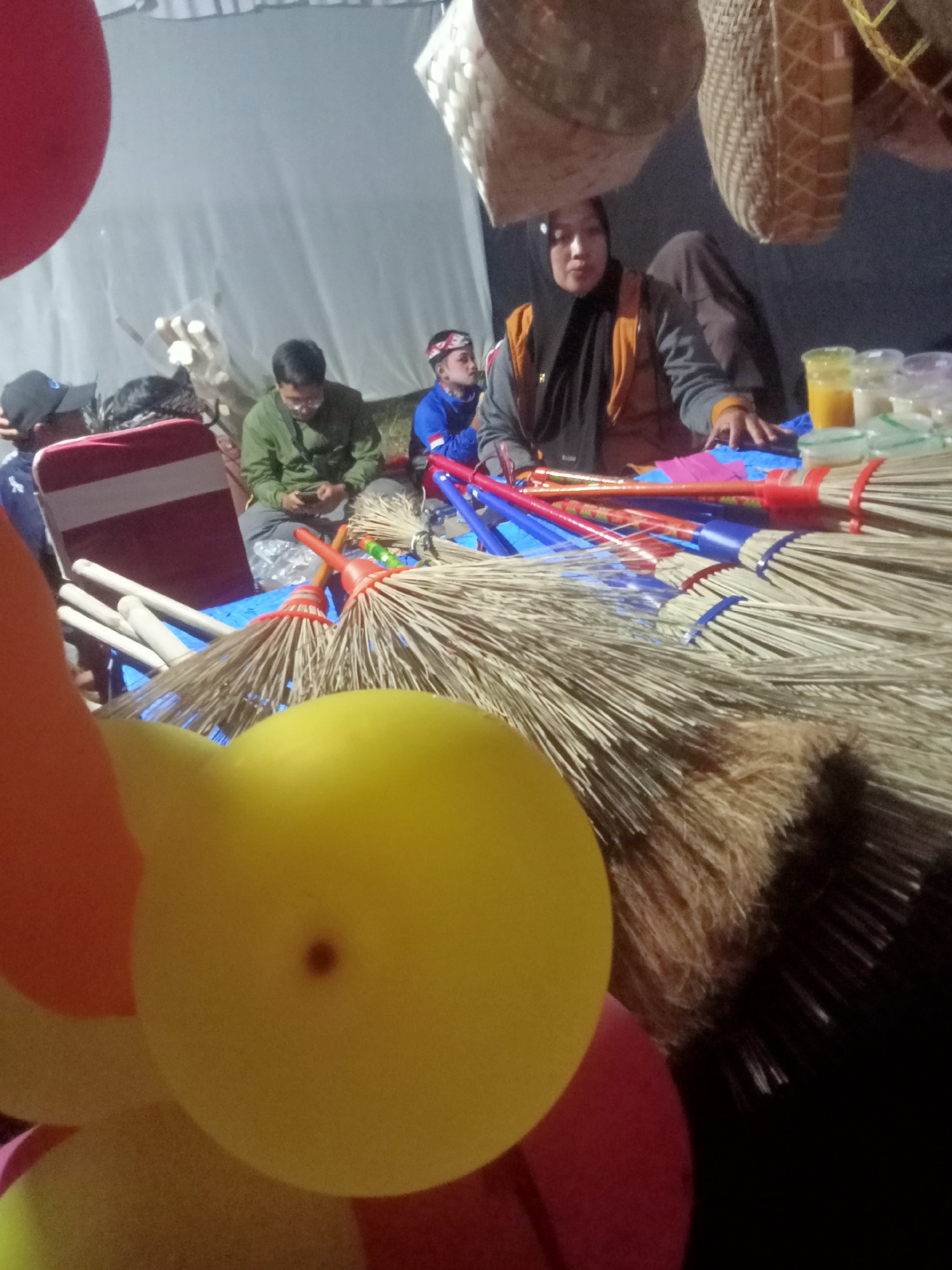 The image depicts an outdoor scene, likely a marketplace or a community gathering, in Pule, Indonesia. In the foreground, several colorful balloons, primarily yellow and orange, are prominently displayed, partially obscuring the view of the background. Behind the balloons, a collection of brooms with natural fiber bristles and colorful handles (red and blue) are arranged.
Further in the background, a woman wearing a black hijab and an orange vest is seated, appearing to be interacting with items on a table. To her left, a man in a green long-sleeved shirt is seated, looking down at his phone, suggesting a moment of pause or interaction with technology. Another individual, possibly a young boy, is visible wearing a blue top with a headband. The setting appears to be under a large white tent or canopy. Woven baskets hang from above, adding to the market-like atmosphere. The overall lighting suggests it might be evening or indoors with artificial lighting.