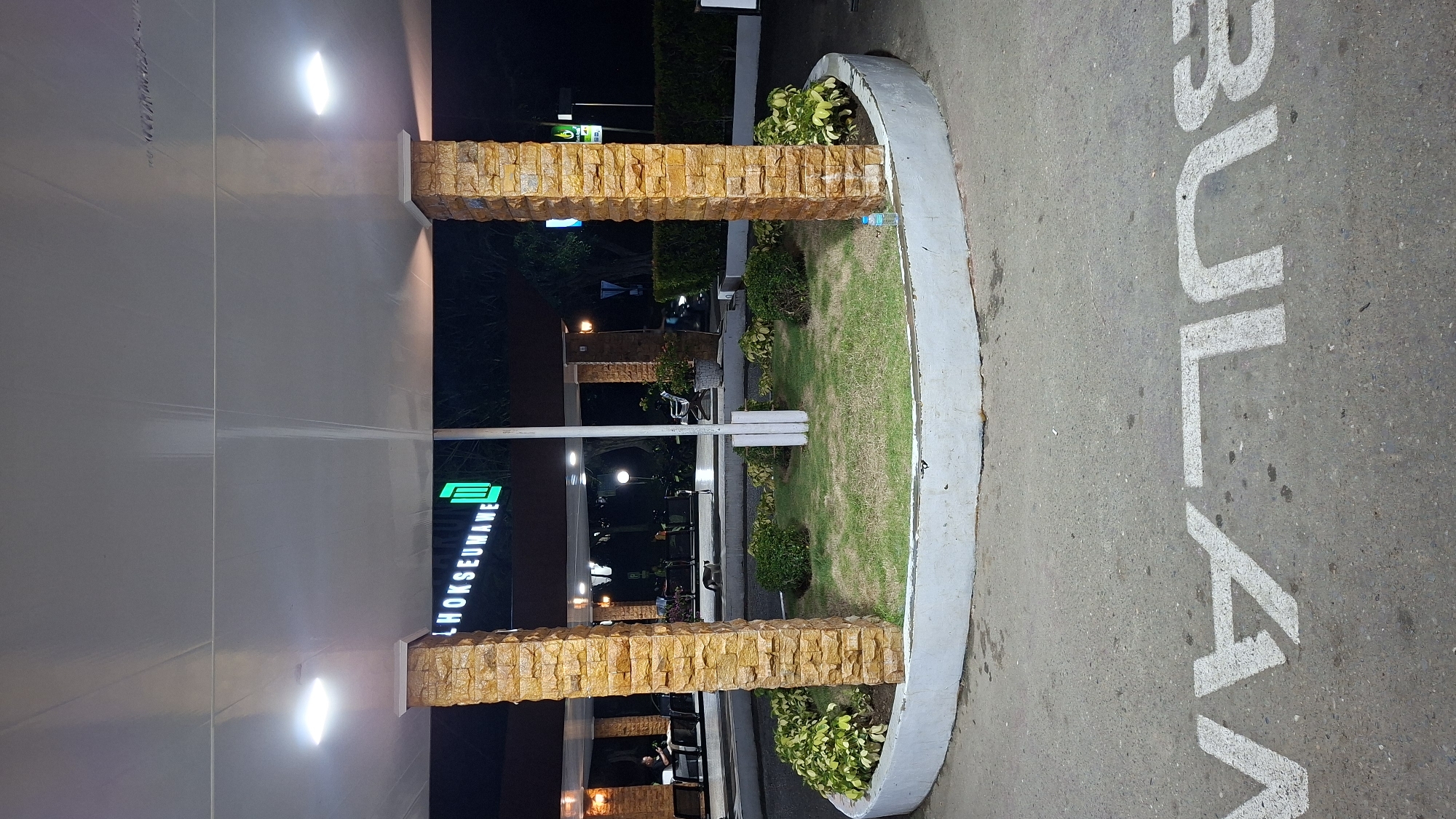 The image depicts a nighttime scene at what appears to be a transportation hub or rest stop in Lhokseumawe, Indonesia. The foreground shows a circular grassy area with small bushes and decorative plants, bordered by a concrete curb. White painted letters on the asphalt road read "BULAN" (moon in Indonesian). Two prominent brick pillars support an overhang. In the background, there is a sign with green lettering that reads "LHOKSEUMAWE" and a stylized "M" logo. Several people are seated at tables under the shelter, with some seated indoors and others outdoors. Artificial lights illuminate the area. The overall impression is of a well-lit public space, likely a service area for travelers.