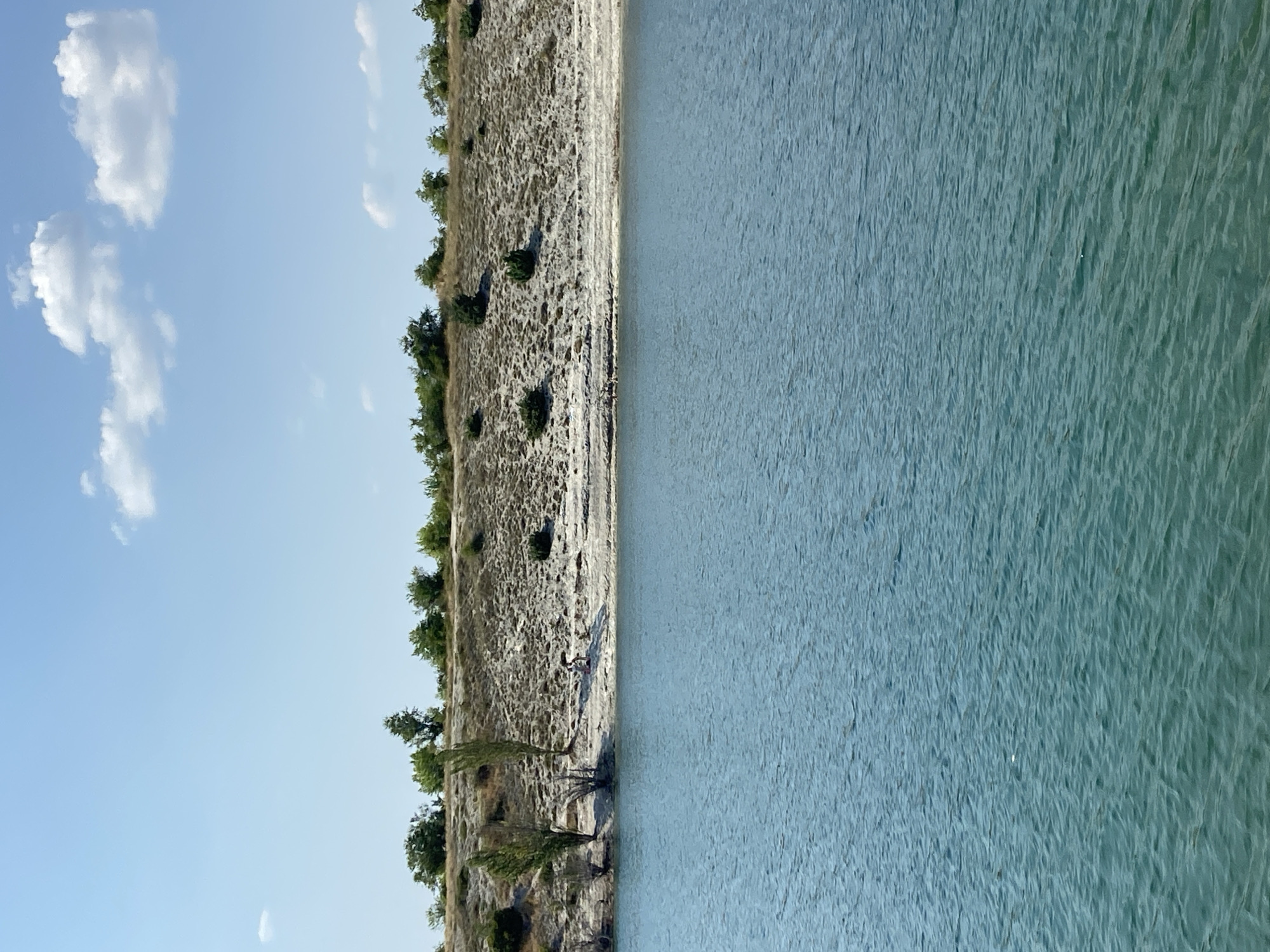 The image shows a view of a body of water with a sandy embankment and vegetation in the background. The water is a turquoise color and has ripples on its surface, suggesting a gentle breeze. The embankment is covered with sparse, dry grasses and scattered bushes, with a few trees growing along the top. The sky is a clear blue with a few white clouds. In the middle ground, on the embankment near the water's edge, two people are visible. One appears to be sitting, and the other is standing. The scene is likely a natural landscape, possibly a lake or reservoir, in or near Куйбышевское сельское поселение, Ukraine.