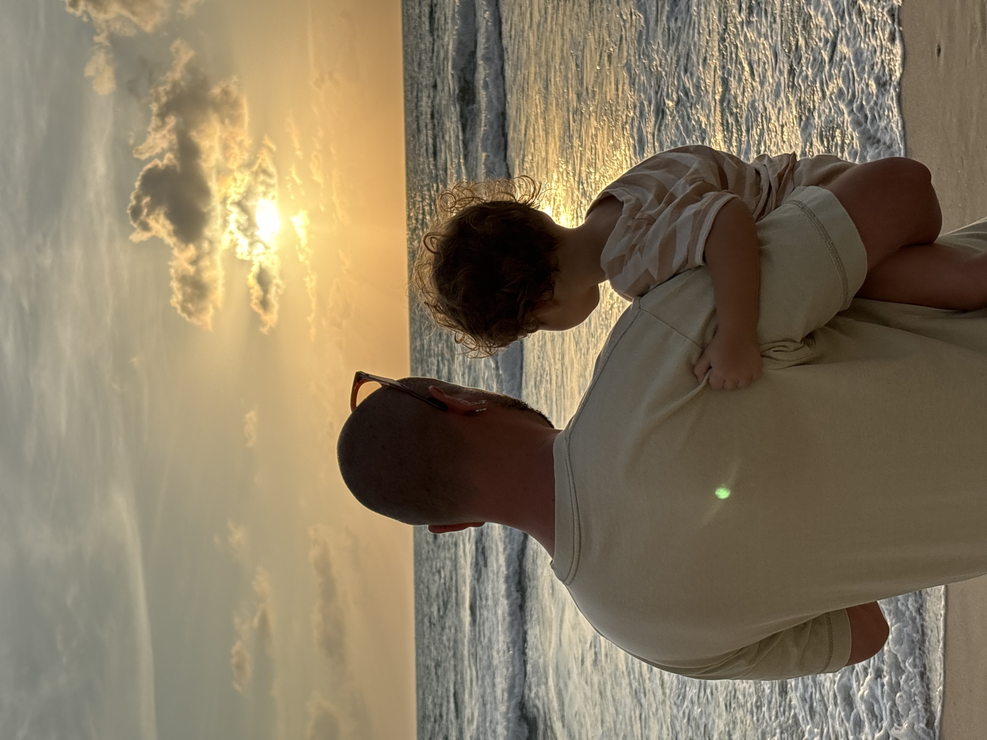 A person holds a toddler on a beach in Choeng Thale, Thailand, during sunset. The person, facing away from the camera, is wearing a light-colored t-shirt and sunglasses pushed back on their head. The toddler, also facing away, is wearing a striped t-shirt and shorts. They are standing on a sandy beach with the ocean in front of them. The sun is setting over the ocean, casting a warm glow on the scene. The waves are gently rolling onto the shore.