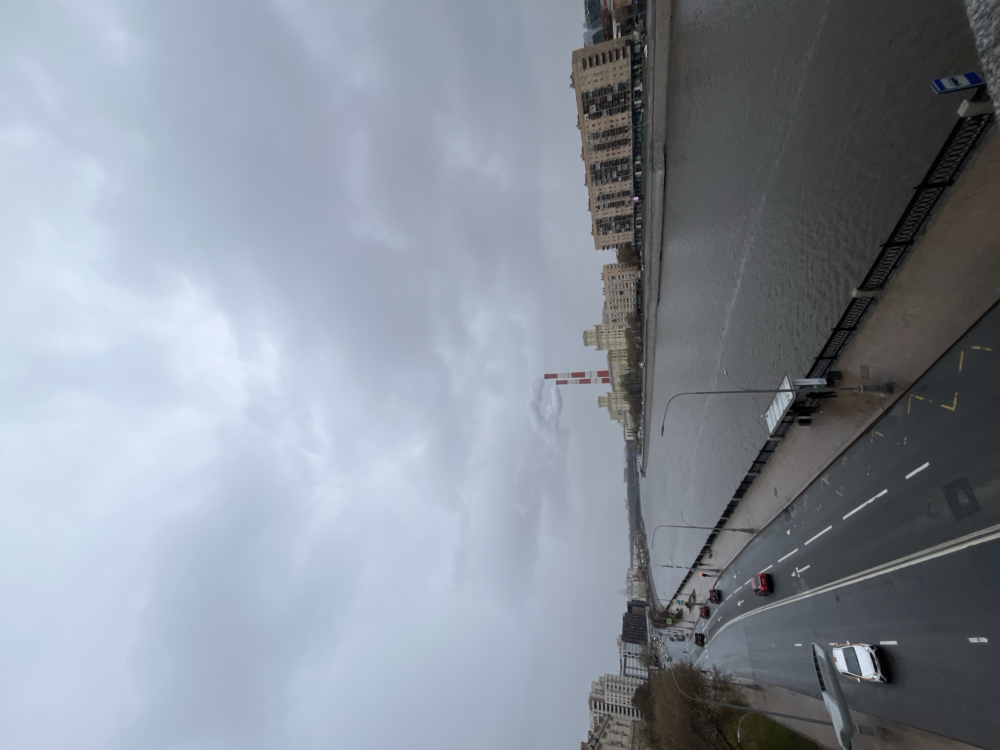 The image captures an overcast scene in Moscow, Russia, featuring the Moskva River and adjacent urban infrastructure. The river flows from the upper right to the middle ground. Along the right bank, a gray concrete embankment with a dark railing runs parallel to a wet asphalt road with visible white lane markings and sparse vehicular traffic. The opposite bank is lined with apartment buildings and other structures. A smokestack, painted with alternating red and white stripes, rises into the heavy sky and releases what appears to be water vapor or light smoke. The sky is a uniform gray, indicating possible rainfall or a heavily overcast day. The overall scene is devoid of any apparent pedestrian activity or human subjects on the visible portions of the embankment or riverside.