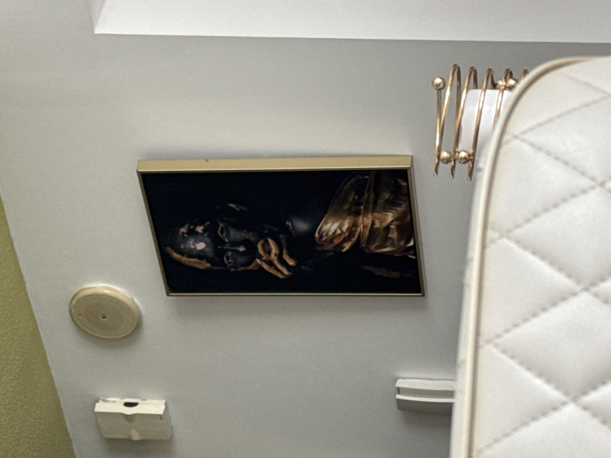 The image depicts an indoor scene, likely a bedroom or living area, with a modern aesthetic. On a light gray wall, a framed artwork features a dark portrait of a person with gold accents on their face and draped clothing, set against a black background. To the left of the artwork, a white electrical box is mounted on the wall, and a circular beige device, possibly a speaker or a sensor, is positioned above and to the left of the artwork. In the foreground, the top portion of a white, quilted headboard or sofa is visible. To the right of the artwork, a decorative golden spiral object with spherical accents is placed on the edge of the furniture. The ceiling in the upper left corner has a textured, light green finish. The overall ambiance is one of contemporary interior design, with a focus on art and decorative elements. The location is not specified, and therefore it cannot be determined if this scene is in Dubai, United Arab Emirates.