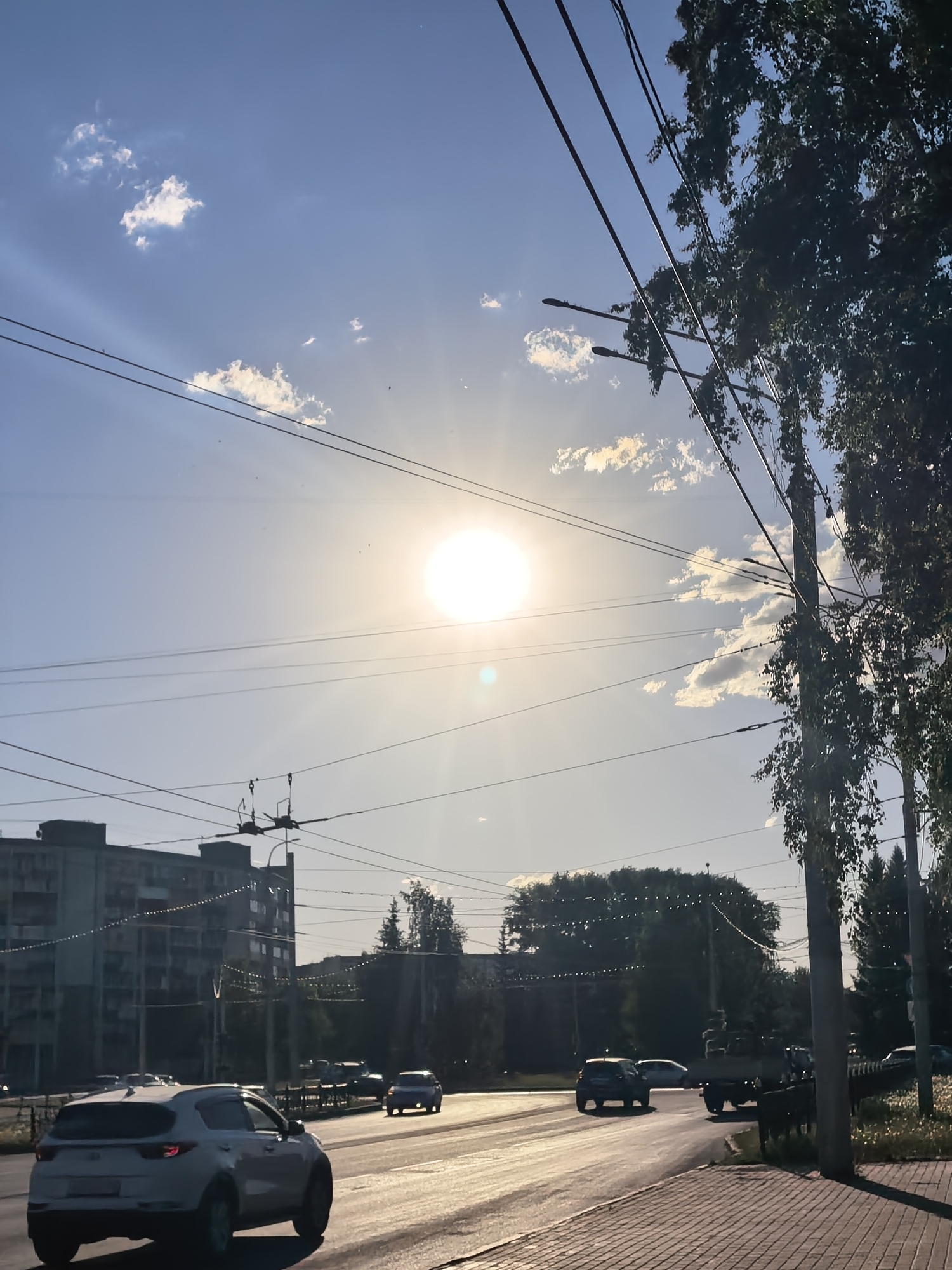 The image shows a street scene in Sterlitamak, Russia on a sunny day. The sun is high in the sky, partially obscured by thin clouds. Power lines crisscross the sky. A multi-story building is visible in the background on the left. A line of trees is on the right. Cars are driving on the road. The road is paved with asphalt, and the sidewalk is paved with brick. The overall lighting is bright.