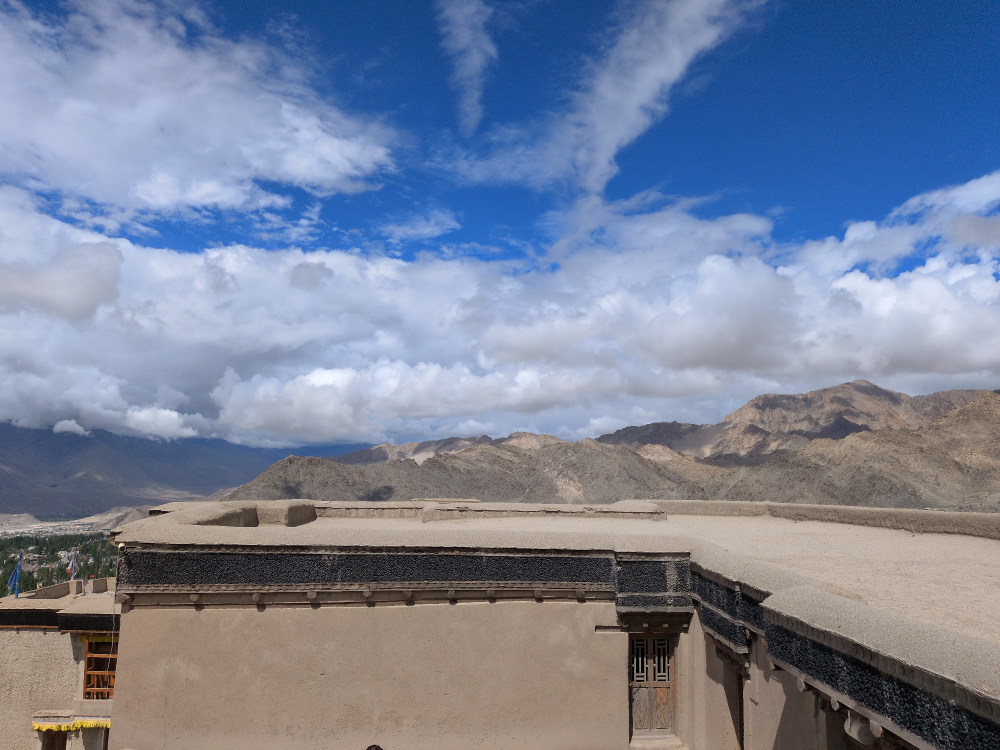 The image captures a view from a rooftop in Leh, India, overlooking a valley and mountainous terrain under a bright blue sky dotted with white clouds. In the foreground, a tan-colored building with intricate dark decorative elements along its parapet dominates the lower portion of the frame. A wooden window with a lattice design is visible on the right side of the building. Beyond the building, a town with scattered trees stretches towards the base of barren, tan-colored mountains. The landscape suggests a high-altitude, arid environment, characteristic of Ladakh.