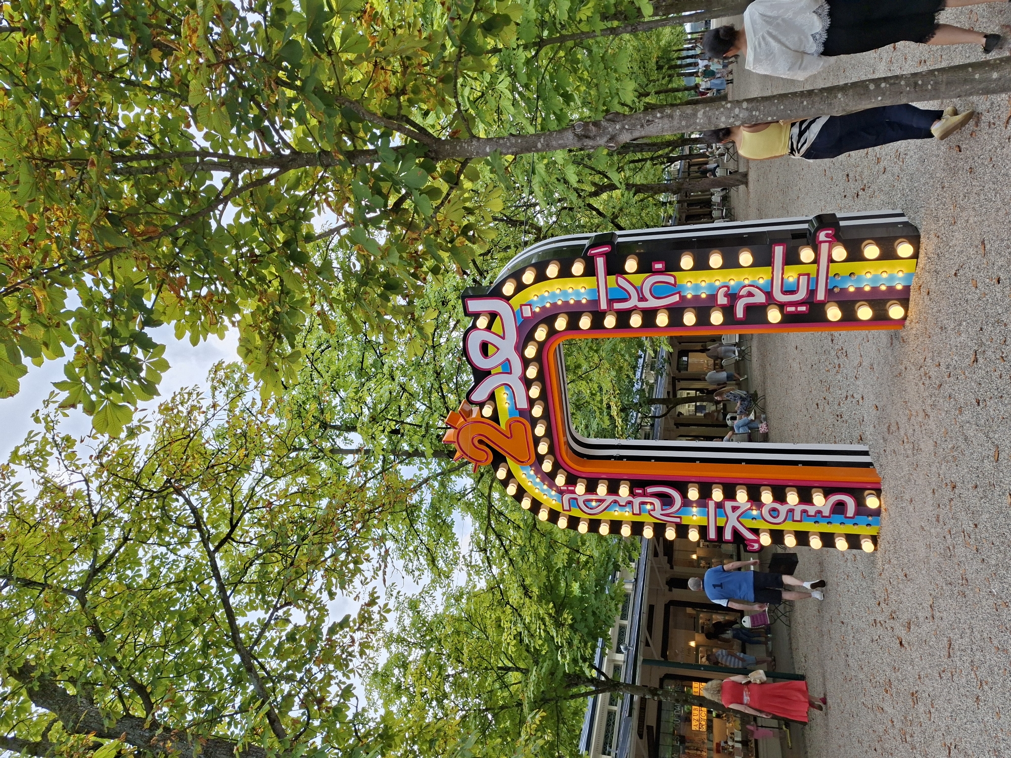 In Baden-Baden, Germany, a vibrant, illuminated archway stands prominently in a park-like setting. The archway, decorated with colorful stripes and numerous light bulbs, features Arabic script and the number "2". The ground is covered with gravel, and trees with green foliage line the path. In the background, a row of buildings with awnings suggests a commercial or recreational area, with tables and chairs visible. Several people are present: a woman in a red dress walks away from the camera, a man in a blue shirt and shorts walks towards the archway, and two women are seen from behind walking away from the archway. Other individuals are seated or walking in the distance, contributing to a lively atmosphere. The lighting and the presence of people indicate this is likely a daytime scene, possibly during an event or festival.