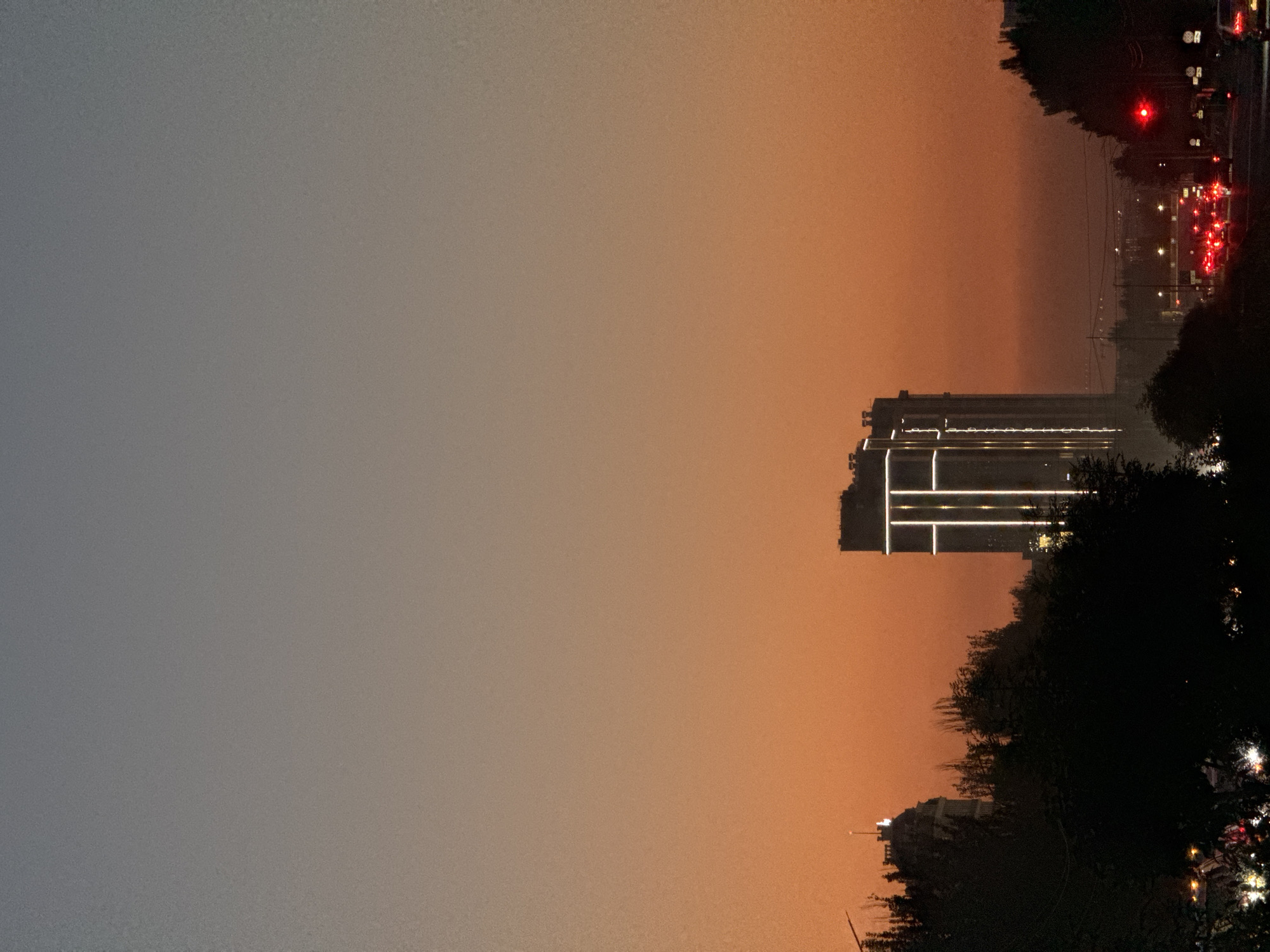 The image depicts a dusky sky over Bishkek City, Kyrgyzstan. A tall, modern building with illuminated vertical lines is prominently featured against the orange and gray gradient of the twilight sky. In the foreground, silhouetted trees and foliage obscure the lower portion of the scene. A street with traffic, indicated by red brake lights and headlights, is visible towards the bottom right, suggesting an urban setting with ongoing activity despite the fading light. The overall atmosphere is serene and urban.