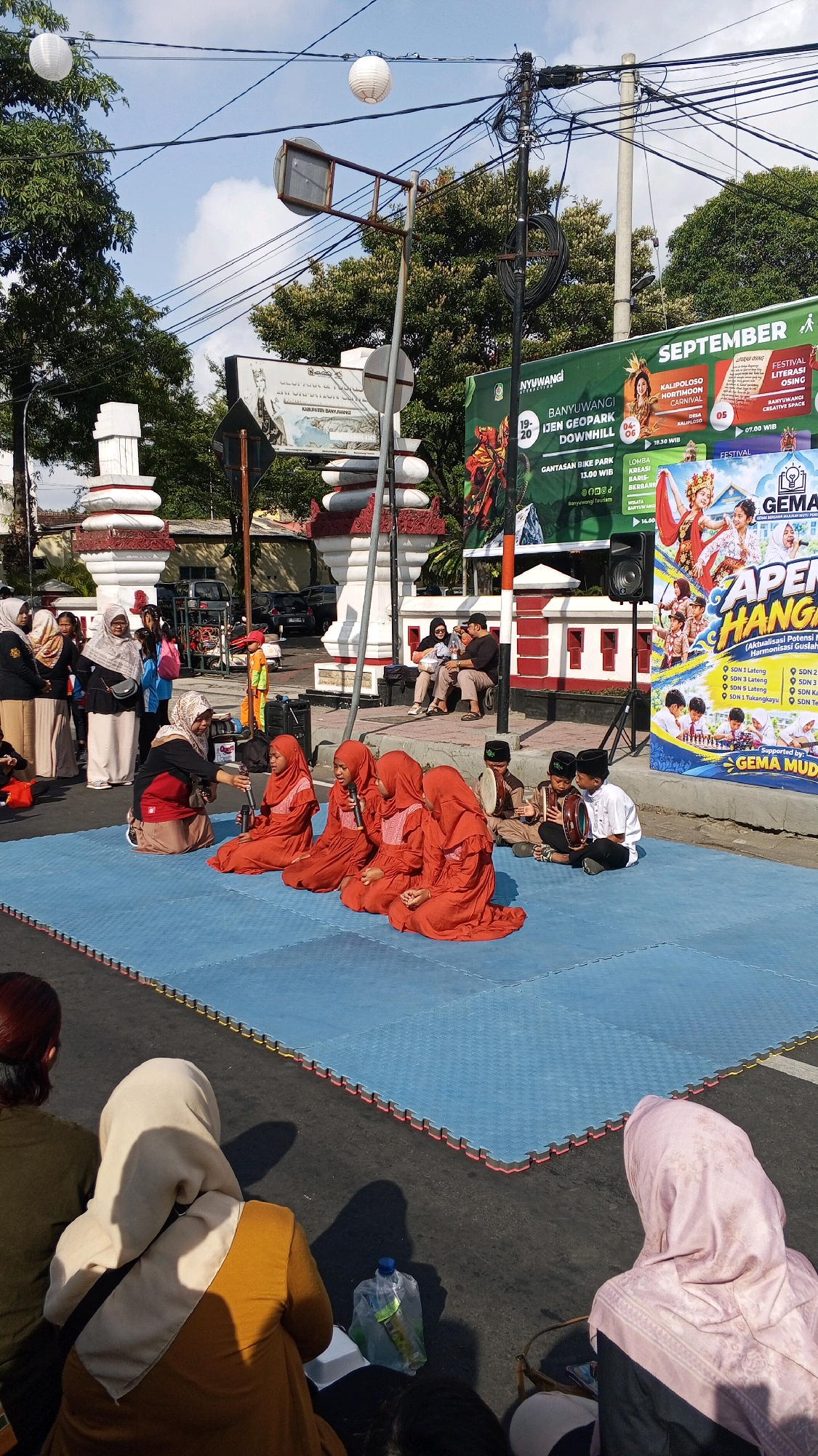 On a sunny day in Tamanbaru, Indonesia, a group of children dressed in vibrant orange hijabs and robes are performing on a blue mat laid on asphalt. Several of the children are holding microphones, suggesting they are singing or reciting. Two boys are seated to the right, playing traditional Indonesian percussion instruments, likely rebana. An adult woman, also in a hijab and brown robe, kneels to the left, facing the performers and holding a microphone. A speaker is set up behind the performers. In the background, onlookers are seated on benches and on the ground, some of them also in hijabs. Beyond the performance area, a large banner with text and images, possibly advertising an event called "GEMA MUDA," is prominently displayed. The street is lined with trees and buildings, and overhead power lines crisscross the sky. Decorative white lanterns are suspended from these lines. The overall scene captures a community event or performance taking place outdoors in a public space.