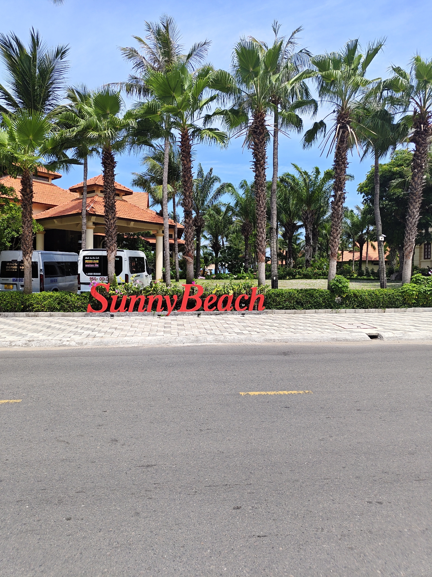 The image depicts a bright, sunny day with a clear blue sky above a row of tall palm trees. In the foreground, a paved road with a yellow painted line runs horizontally across the bottom of the frame. To the left of the road, a sidewalk made of interlocking gray pavers is visible. A large, red, three-dimensional sign that reads "SunnyBeach" is placed on the sidewalk, partially obscured by palm trees. Behind the sign, a well-maintained garden with green bushes and some flowering plants can be seen. Further back, a building with a terracotta-tiled roof and beige walls is visible, with several large columns supporting an overhang. Two white vans are parked in front of the building. The overall scene suggests a tropical resort or hotel entrance.