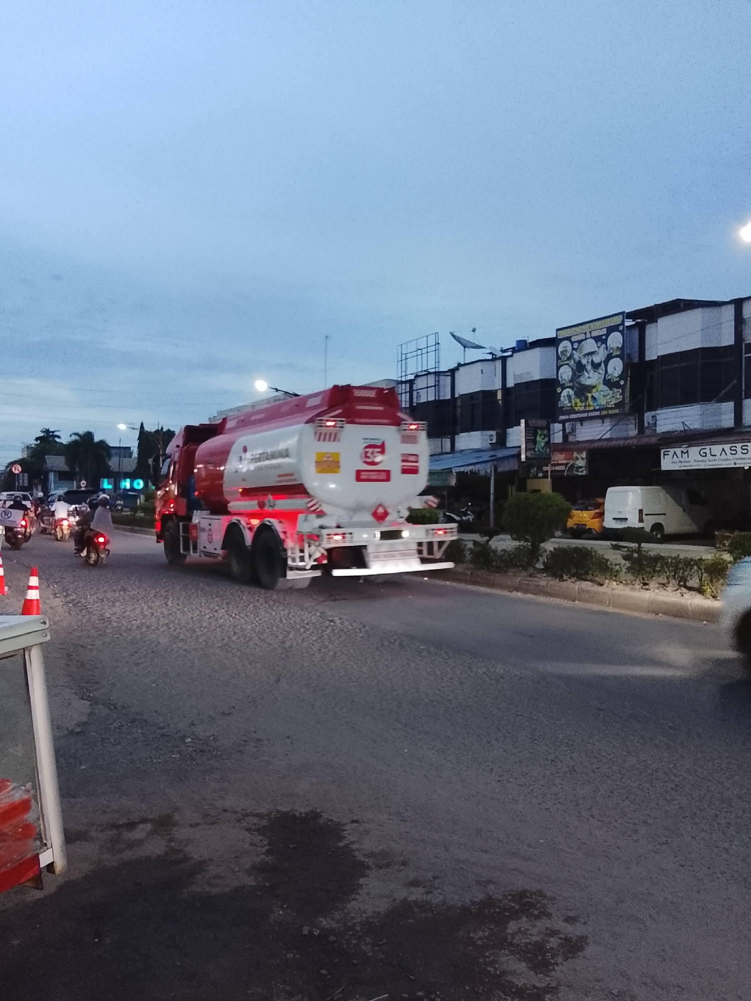In Lhokseumawe, Indonesia, a red and white Pertamina fuel tanker truck is captured in motion on a paved road. The truck, positioned in the center-right of the frame, displays its prominent Pertamina logo. The road is textured with gravel and asphalt, showing some dark patches that could be wet spots. In the background, buildings line the street, one of which features a large banner with images of skulls. Several motorcycles, some with riders, are present on the road, suggesting traffic. Orange traffic cones are visible on the left side of the image. The sky is overcast with a light blue hue, indicating dusk or dawn. A white van is parked in front of the buildings.