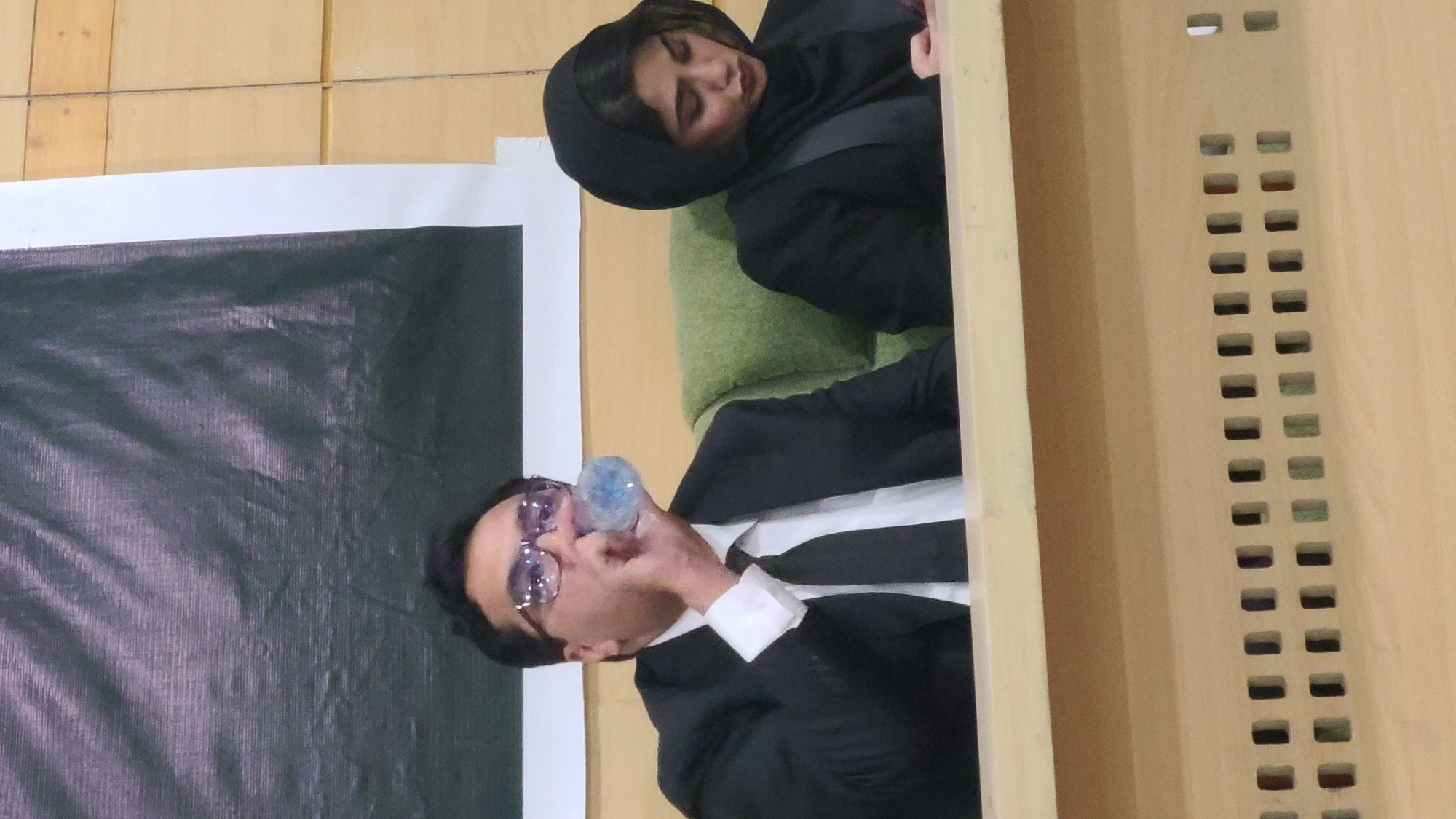 An indoor scene, likely a formal event in Quetta City Tehsil, Pakistan, is captured from a slightly elevated perspective. In the foreground, a man is visible from the chest up, dressed in a black suit, white shirt, and a black tie. He wears distinctive purple-tinted glasses with sparkling accents and is actively drinking from a clear plastic water bottle. His dark hair is neatly styled.
Behind the man and slightly to his right, a woman is seated on what appears to be a green cushioned chair or sofa. She is wearing a black hijab and black attire, looking down with a serene or thoughtful expression.
The background features light-colored wooden paneling, possibly part of a stage or wall. On the right side of the wooden structure, a series of rectangular ventilation-like slits are visible, arranged in two vertical columns. To the left, a large, dark-colored banner or screen, possibly made of fabric, serves as a backdrop. No discernible text is visible on the banner or anywhere else in the image. The lighting suggests an indoor setting, possibly during the daytime. The overall atmosphere appears calm and focused.