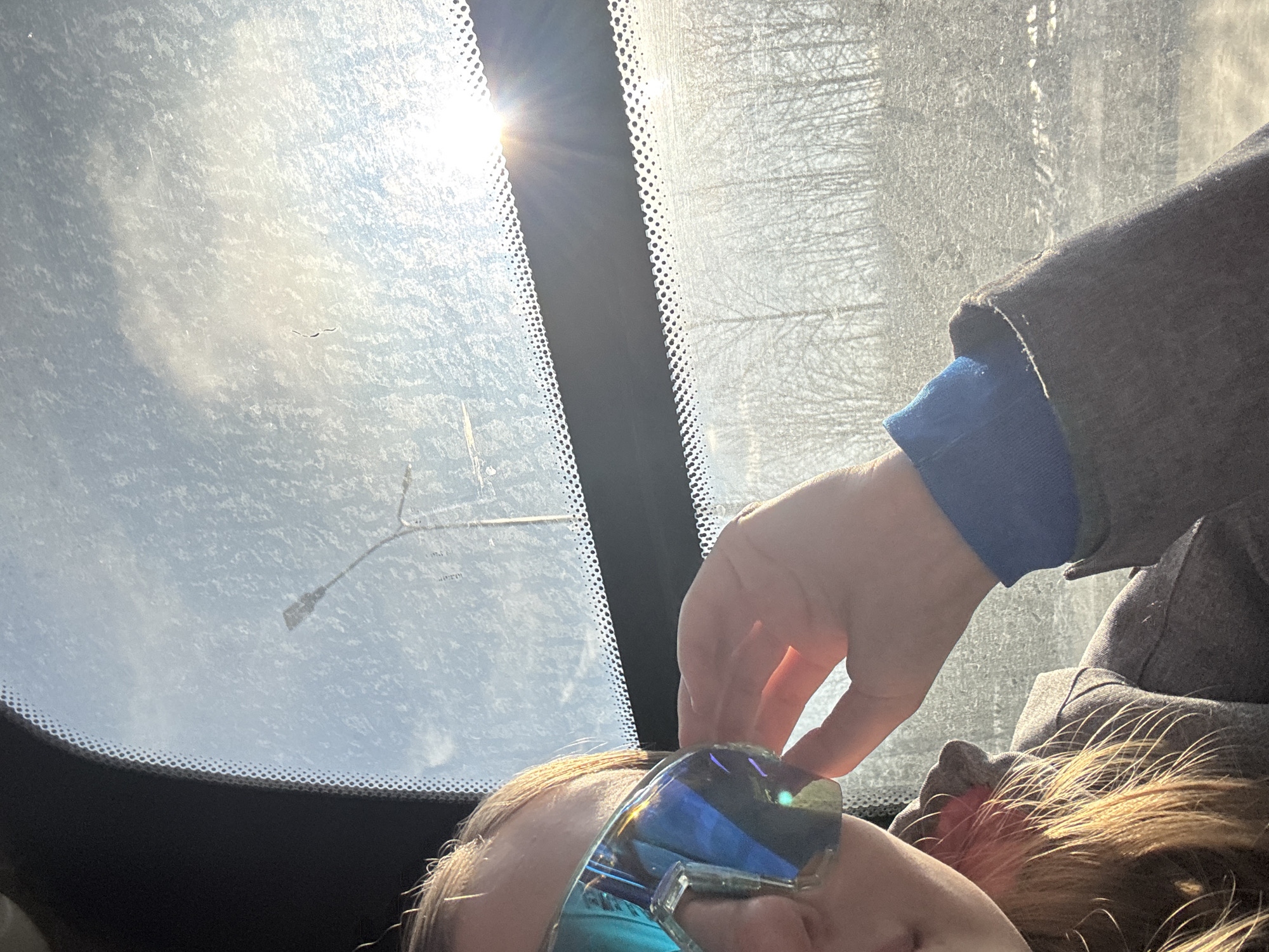 A person, seen from a low angle inside a vehicle, is adjusting or holding sunglasses with a blue reflective lens. The person is wearing a blue shirt underneath a gray jacket. A hand is partially visible near the sunglasses. The vehicle's windshield is visible, displaying a bright sunny sky with a visible street light. The edges of the windshield reveal a patterned black border. Beyond the windshield, trees are faintly visible. While it's impossible to determine the location specifically as Berezniki, Russia from the image alone, the scene shows a sunny outdoor view through a car window.