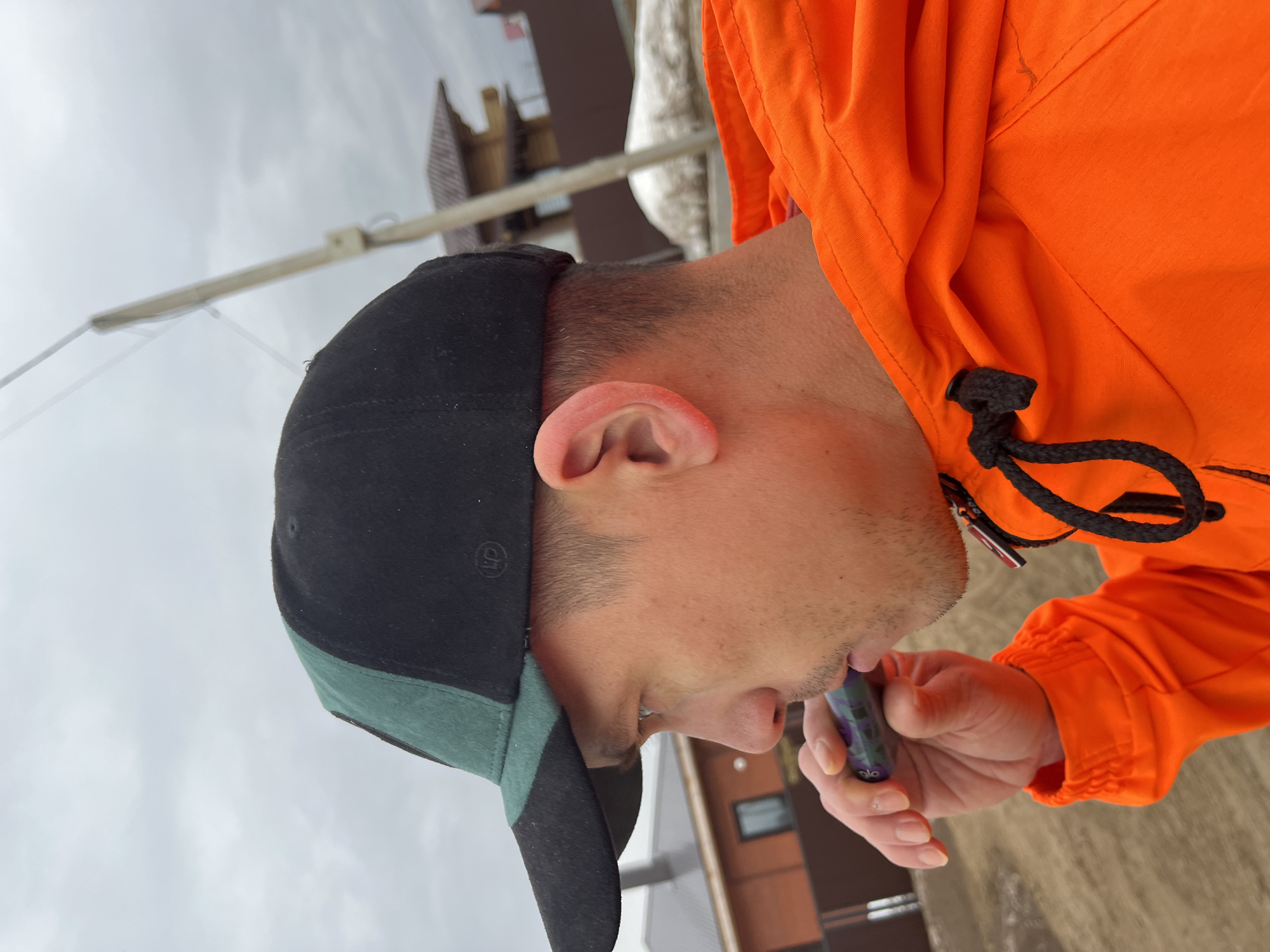 A person is depicted outdoors, holding an electronic cigarette or vaping device to their lips. The individual wears a black and green baseball cap, turned backwards, and an orange hooded jacket. The background shows a building or structure with a window, and a pole extending upwards. The image appears to be taken in Chaikovsky, Russia, given the prompt.