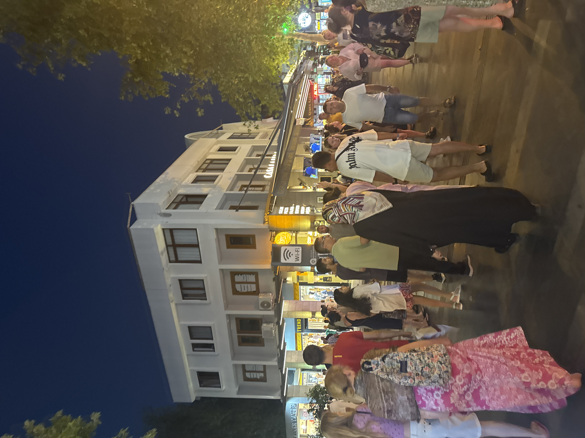 An outdoor nighttime scene depicts a bustling street in Kemer, Turkey, likely a commercial area. A multi-story white building with numerous windows and some air conditioning units dominates the background. Shops with awnings and illuminated displays line the ground floor. A prominent "Wi-Fi" sign is visible on one of the shop fronts. A crowd of people, diverse in age and gender, walks along the paved street, some appearing to be tourists. They are dressed in casual summer clothing. Several individuals are walking towards the camera, while others are walking away. The atmosphere appears lively, possibly indicating a popular evening gathering spot. Trees with foliage are visible on the right side of the frame, with their leaves illuminated by artificial lights. The sky is dark, indicative of nighttime.