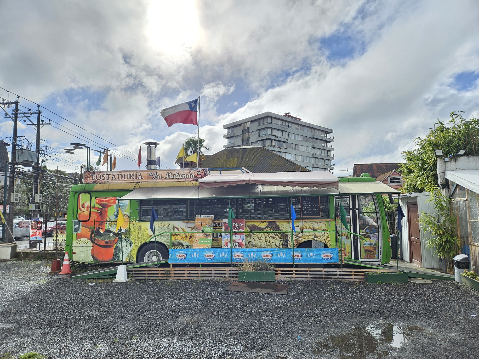 The image showcases a green, food-truck-like structure, emblazoned with the words "TOSTADURÍA 'La Molienda'", located in what appears to be Osorno, Chile, identifiable by the Chilean flag waving atop it. The food truck is adorned with images of various food items, including coffee beans, bread, and sandwiches. The side of the truck advertises a menu featuring items like "CAFÉ-TE", "CAFÉ DE C", "CHOCOLATE", and "LECHE CON MANI", along with "Mote con huesillo", a popular Chilean drink. The truck is elevated on a wooden platform, and a sign featuring the word "Savory" repeats along its base.
The surrounding environment consists of a gravel parking area, with visible signs of moisture, suggesting recent rain. In the background, a modern apartment building can be seen, providing an urban context to the scene. Utility poles and power lines are visible in the periphery. The presence of additional structures, including what appears to be a greenhouse or garden area to the right, suggests the food truck is situated in a mixed urban and semi-rural environment. The sky is partially cloudy. There is no evident people.