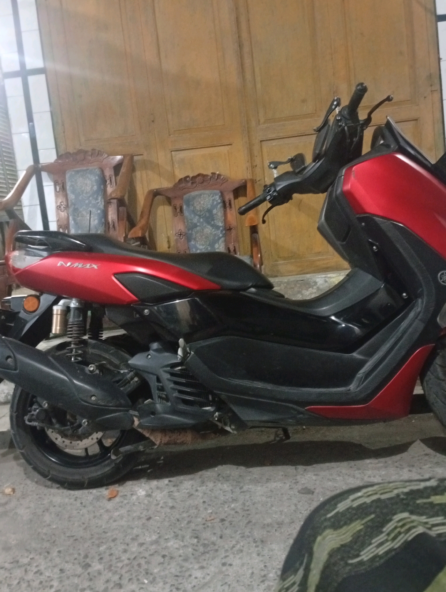 A red and black Yamaha NMAX scooter is parked on a concrete surface. The scooter is facing left and is viewed from the side. The background consists of a wooden door and two ornate wooden chairs with blue upholstery. The scene appears to be an outdoor area, possibly a driveway or courtyard, in Banowan, Indonesia. The lighting is dim, suggesting it may be evening or an overcast day. No people are visible in the image.