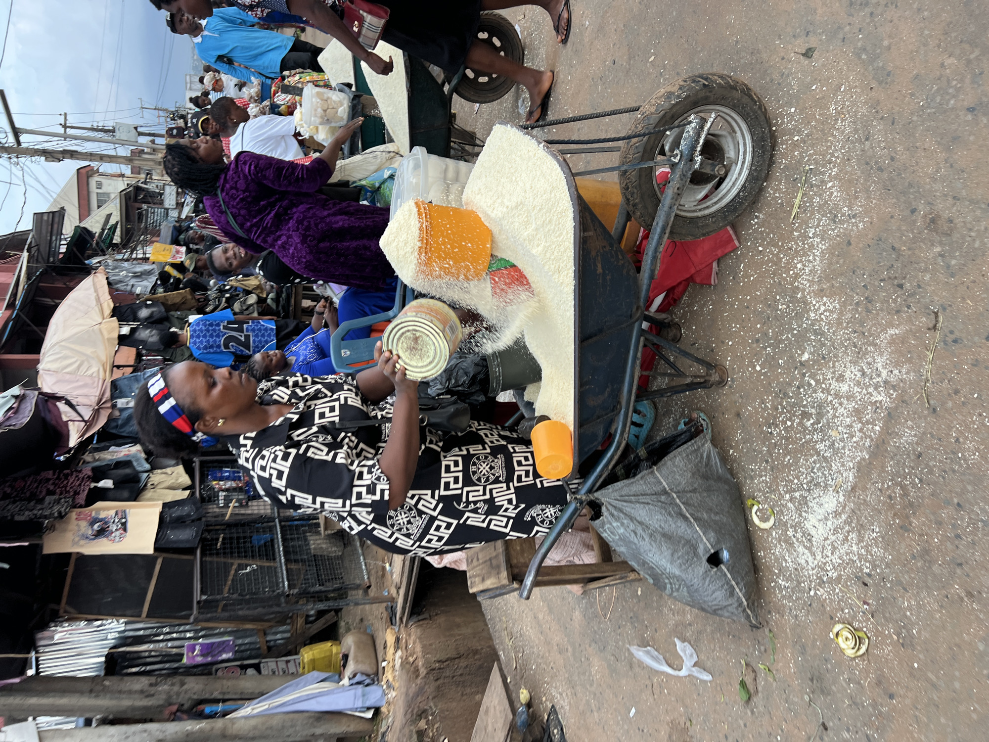In Nkpor, Nigeria, a woman in a black and white patterned dress is at a market stall, transferring rice from a metal can into an orange plastic bucket that sits atop a wheelbarrow overflowing with rice. She wears a red, white, and blue headband. The wheelbarrow is laden with a large quantity of white rice, which spills over the sides. Another wheelbarrow filled with rice is visible to the right, behind a person in a purple garment. The market appears to be outdoors, with various stalls and goods, including clothing and other merchandise, lining the street. A large black plastic bag lies on the ground near the wheelbarrow. The ground is a mixture of dirt and concrete, and scattered grains of rice are visible on the surface. Several other individuals are present in the background, engaged in market activities. The overall scene depicts a busy marketplace in an urban Nigerian setting.