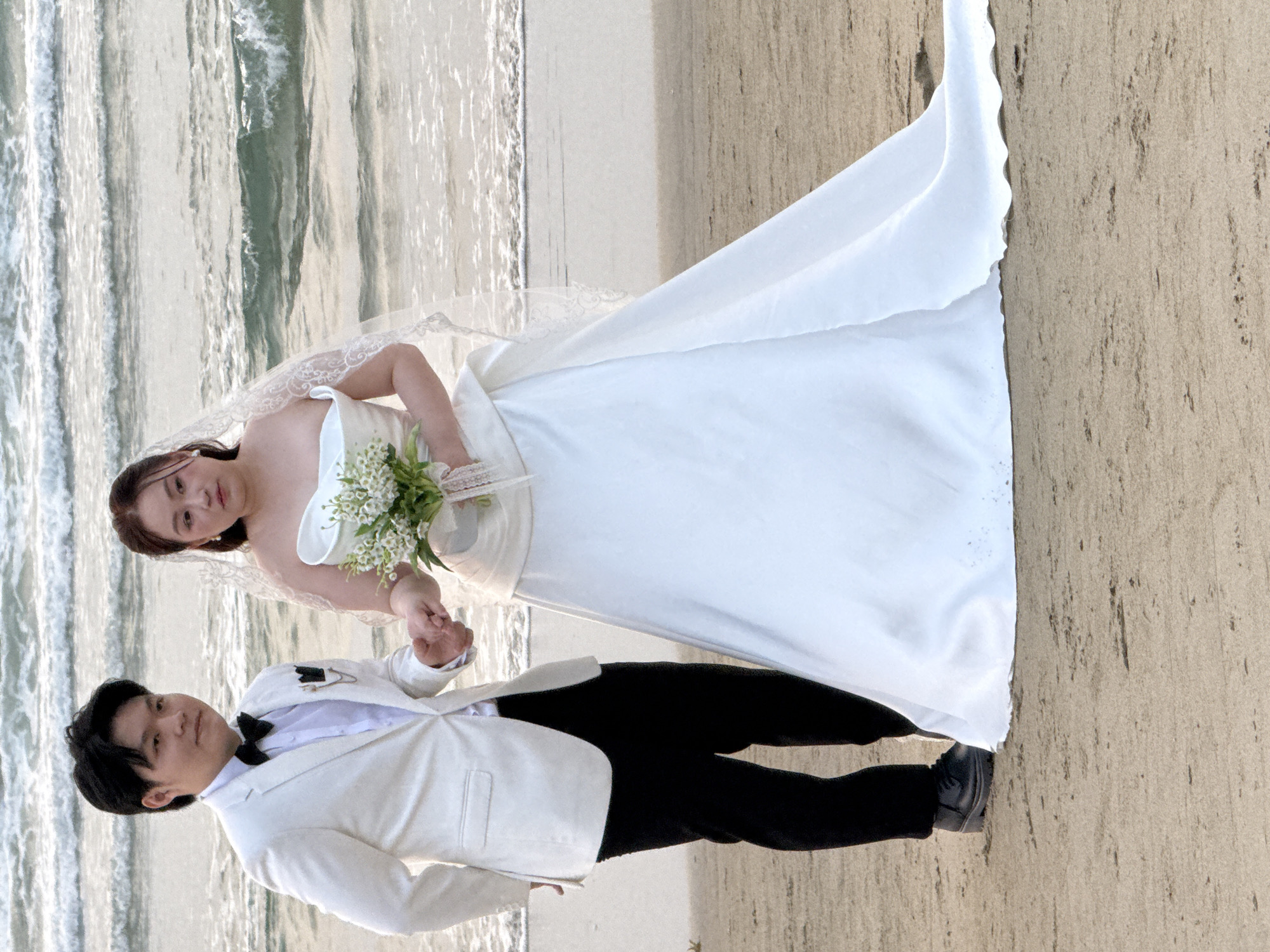 A wedding photo shows a couple standing on a beach. The bride wears a white dress with a veil and holds a bouquet of white flowers. The groom wears a white jacket and black pants with a bowtie. The background includes the ocean and the beach. Based on the appearance of the beach and ocean, the location may be in Đà Nẵng, Vietnam.