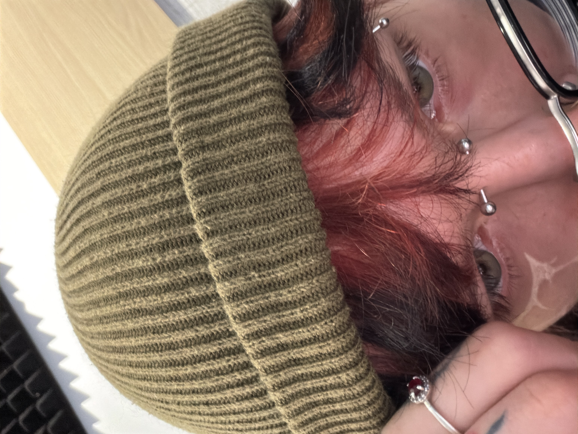 A close-up, slightly rotated image shows a person wearing a ribbed, olive green beanie. The person has short, reddish-brown hair peeking out from under the beanie. They have multiple piercings visible on their face, including piercings above and below their eyes. They are wearing glasses. A hand is visible near the bottom right of the frame, with a silver ring that has a dark red stone. The background appears to be a light-colored wooden surface with some light and shadow patterns from an unknown source. There is no discernible information to identify the location as Sofia, Bulgaria.