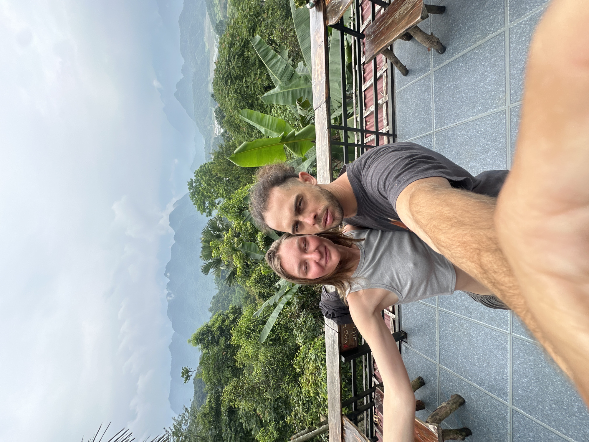 A man and woman take a selfie on a balcony overlooking a lush green valley with mountains in the background, under a cloudy sky. The woman, on the left, wears a grey tank top and a necklace. The man, on the right, wears a dark t-shirt. They are standing in front of wooden tables and benches on the balcony, which has a grey tiled floor and a metal railing. Banana trees and other tropical foliage are visible in the middle ground, with layers of mountains receding into the distance. The overall scene suggests a viewpoint in Pù Luông Commune, Vietnam, known for its natural beauty.