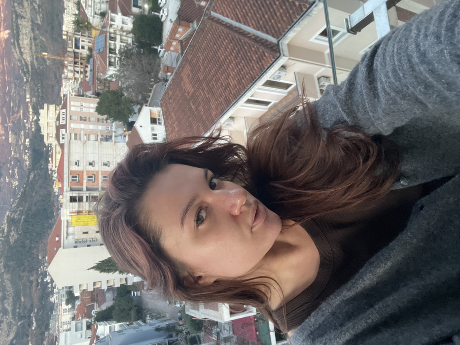 The image displays an adult female in a selfie-style shot, positioned in the foreground and appearing to be on a balcony overlooking the city of Budva, Montenegro. She has long, wavy dark brown hair and is wearing a dark brown scoop-neck top beneath an open, light gray knitted cardigan. Her expression is neutral with slightly pursed lips, and her gaze is directed toward the camera.
The background presents a panoramic view of Budva, characterized by a dense arrangement of multi-story buildings, predominantly white or light-colored, with numerous terracotta-tiled roofs. Evidence of ongoing construction includes a building partially covered with yellow material and scaffolding, and a visible construction crane in the mid-ground. Several buildings feature externally mounted air conditioning units. A street with multiple parked vehicles is visible below and to the left of the main subject. The distant background features rolling, vegetated mountains under a clear sky, illuminated by soft, warm light suggesting either sunrise or sunset. A glass balcony railing is partially visible in the lower right portion of the frame.