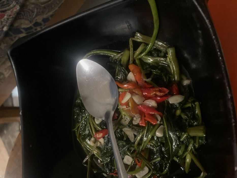 A black, square-shaped serving bowl contains a prepared dish of dark green, stir-fried water spinach (kangkung), visibly intermingled with numerous sliced red chili peppers and chopped white garlic cloves. A single silver-colored metal spoon lies horizontally across the top surface of the food. The background is blurred, revealing a light brown, possibly wooden, surface in the upper left, a patterned textile in the upper right, and a solid orange surface in the lower right, consistent with an indoor dining environment. This scene is located in Cimenyan, Indonesia.