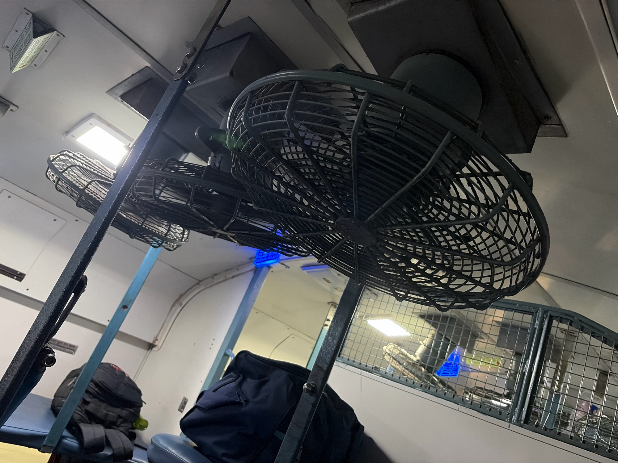 The image depicts the interior of a train compartment, likely in India, given the typical overhead fans and seating arrangement. Two dark-colored, oscillating fans are mounted on the ceiling. The fan in the foreground is larger and closer to the camera, with its wire cage clearly visible. Another fan, smaller and further back, is partially obscured. In the lower left of the frame, a dark backpack rests on a blue seat. To the right, a dark blue garment, possibly a jacket or bag, is placed on another seat. The walls of the compartment are off-white. Fluorescent lights are visible on the ceiling, illuminating the space. A metal grid structure is visible in the background, characteristic of train compartments. There are no people clearly visible in the image. The overall impression is a utilitarian train interior, with luggage suggesting recent or upcoming travel. The image does not provide any specific details about the city of Dewas, India, but the context of a train compartment is consistent with travel within India.