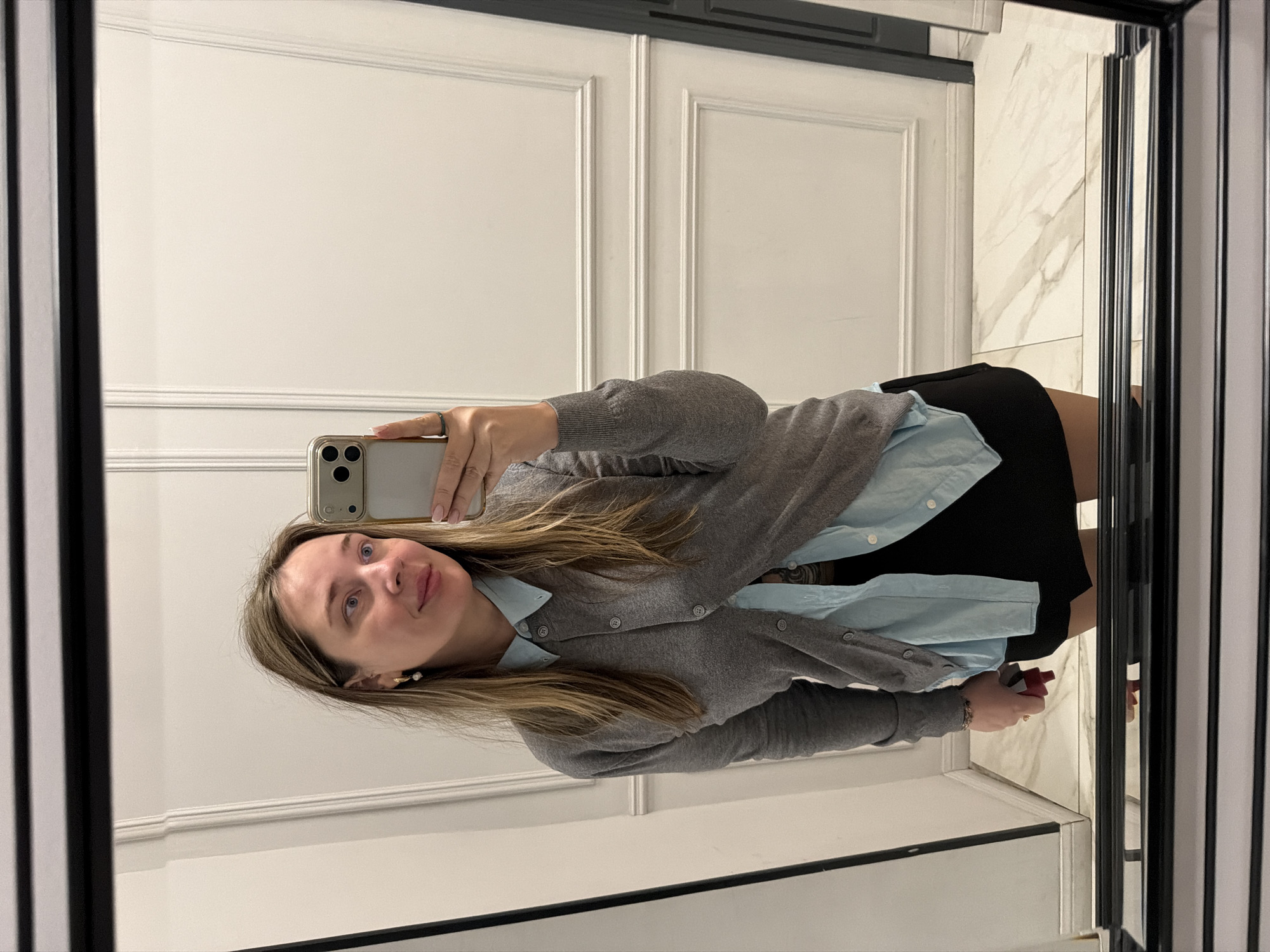 A woman is taking a selfie in a mirror. She is wearing a light blue button-down shirt, a grey cardigan, and a black skirt. Her hair is blonde and falls to her shoulders. She is holding a smartphone in her left hand to take the picture.
The background is a wall with white, paneled doors. To the right of the woman, there is a narrow strip of white marble with grey veins. The mirror has a black frame.
The location cannot be determined from the image. The city of Moscow, Russia, cannot be confirmed.