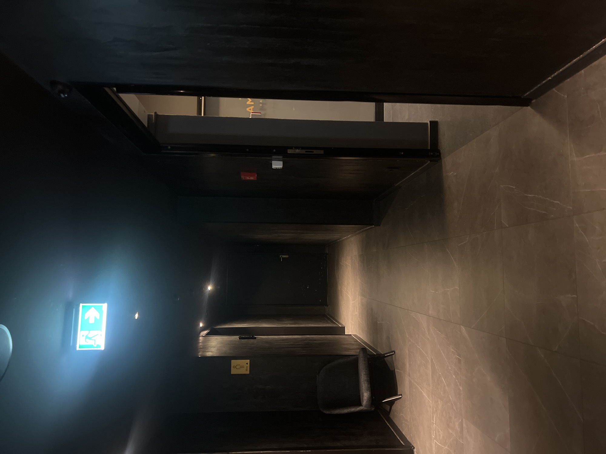 The image depicts a dimly lit interior hallway with dark walls and marble-tiled flooring. An illuminated green exit sign with a running man icon is visible on the ceiling. To the left, a single dark chair is positioned against the wall. Further down the hallway, a door is visible on the right side, and another on the left. On the left wall, a sign with a male icon and the word "Men" is affixed. The hallway appears to lead to an open doorway on the right, revealing a partially visible interior with frosted glass and text that appears to read "HAM" and "EAT." The scene is quiet and still, with no people visible. The overall impression is of a modern, minimalist interior, possibly a hotel, spa, or entertainment venue, located in Berlin, Germany.