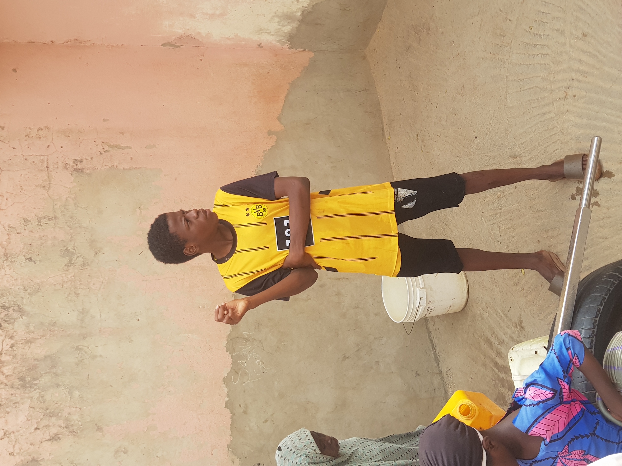 A young man wearing a yellow and black soccer jersey stands in the corner of a room or courtyard in Dutsinma B, Nigeria. The walls are painted in shades of pink and tan, and show signs of age and wear. The ground is a similar tan color. Near the man's feet are a white bucket and a tire leaning against a metal object, possibly a stand or tool. To the left of the tire, partially obscured, are figures lying on the ground; one person is covered in a blue and pink patterned fabric and another in a light-colored, patterned headscarf. A yellow container is also visible. The overall scene suggests a domestic setting or an informal gathering.