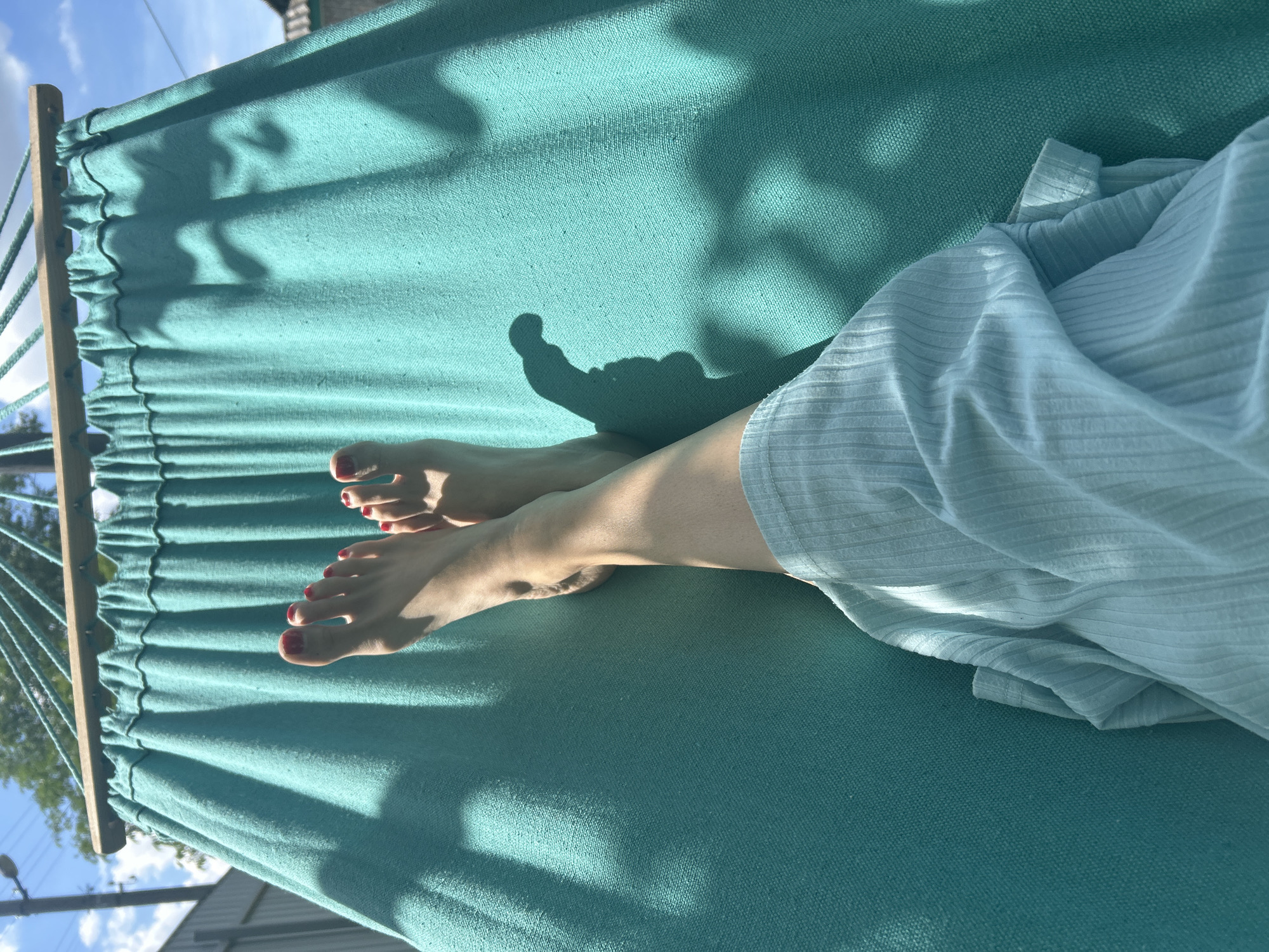 The image shows a close-up view of a person's legs and feet lying in a teal-colored hammock. The person's feet are bare, with toenails painted red. They are wearing a light blue, ribbed garment that drapes over their legs. Sunlight casts shadows on the hammock, suggesting it is daytime, likely outdoors. The background is blurred but shows a glimpse of blue sky with white clouds, trees, and what appears to be a building or fence. The overall impression is one of relaxation and leisure. There is no visible text in the image. The context provided indicates the location is Vyshevychi, Ukraine.